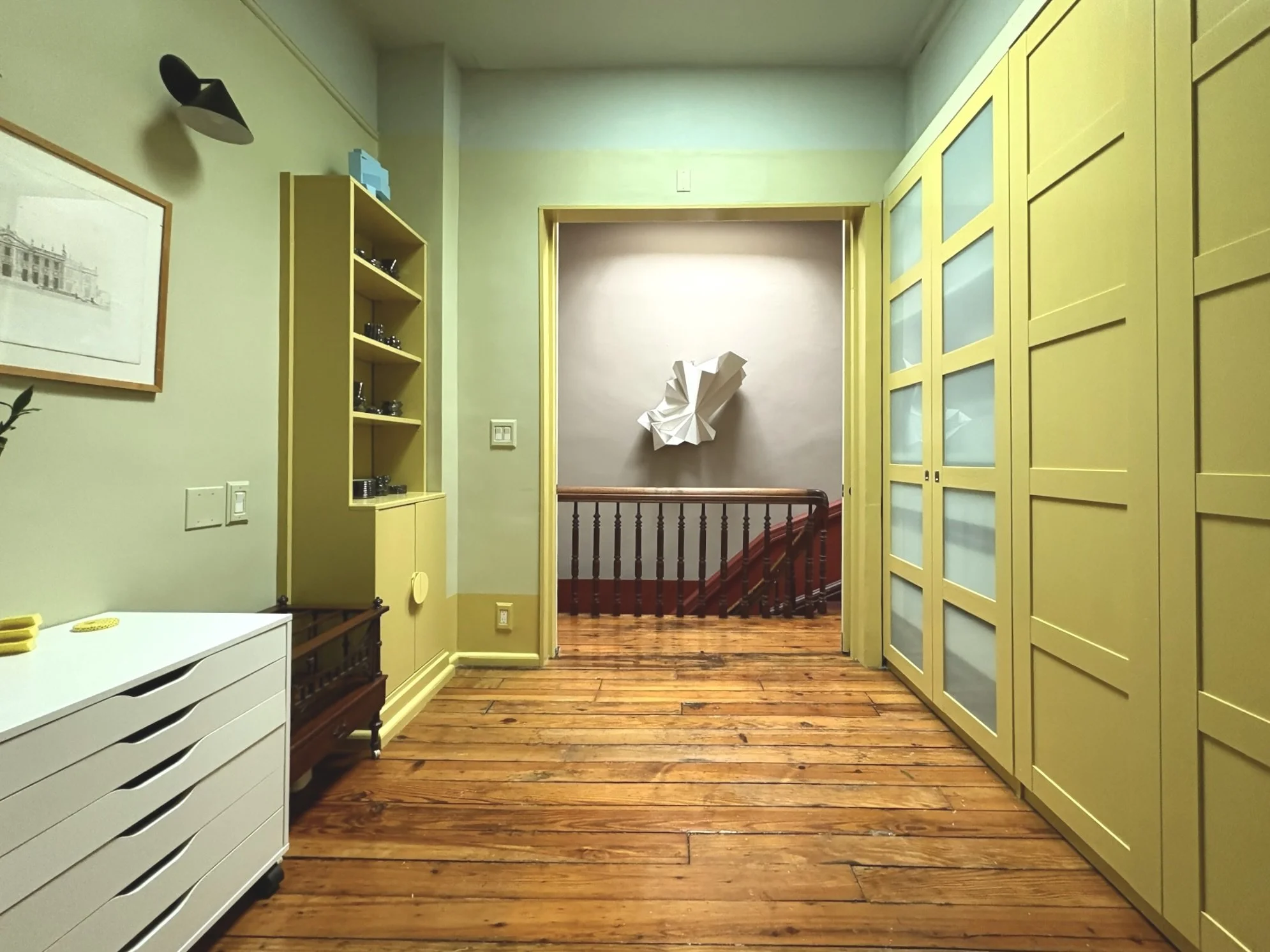 Interior of a room with yellow cabinets, a white dresser with yellow items on top, a black wall lamp, wood flooring, and a wall decoration at the end of the hallway leading to a staircase.