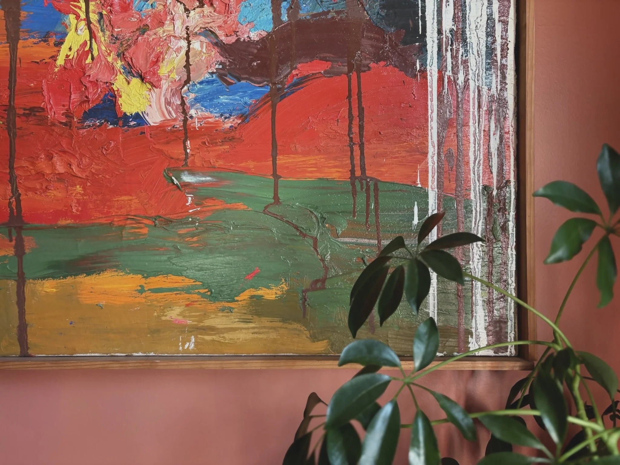 Abstract landscape painting with vibrant red, blue, yellow, and green colors, framed and displayed on a pink wall, with a green leafy plant partially covering the bottom right corner.