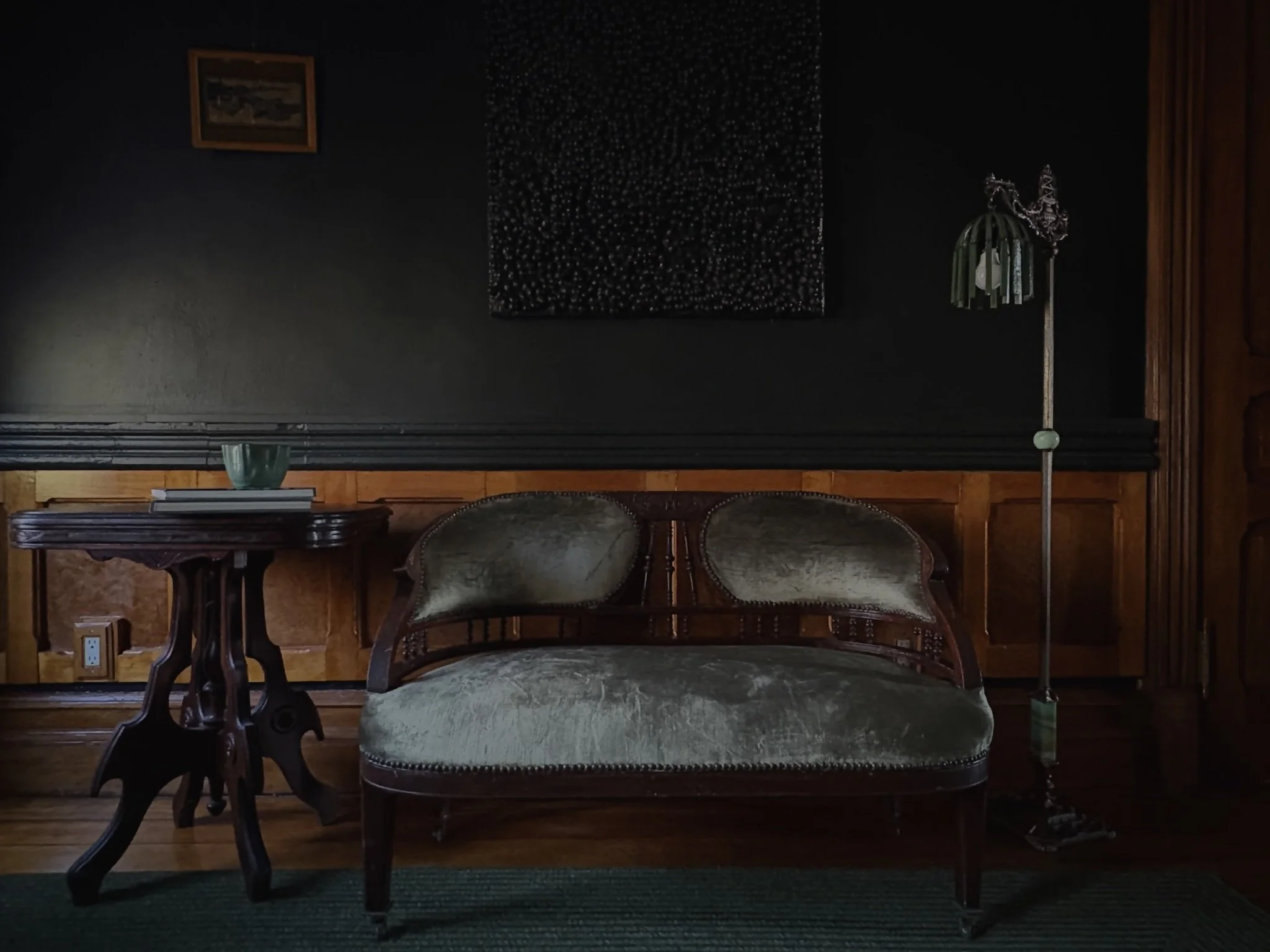 Vintage wooden loveseat with padded velvet cushions, next to a small carved wooden side table with a green bowl and books on it, set against a dark wall with wooden paneling, part of a floor lamp with a green shade to the right.