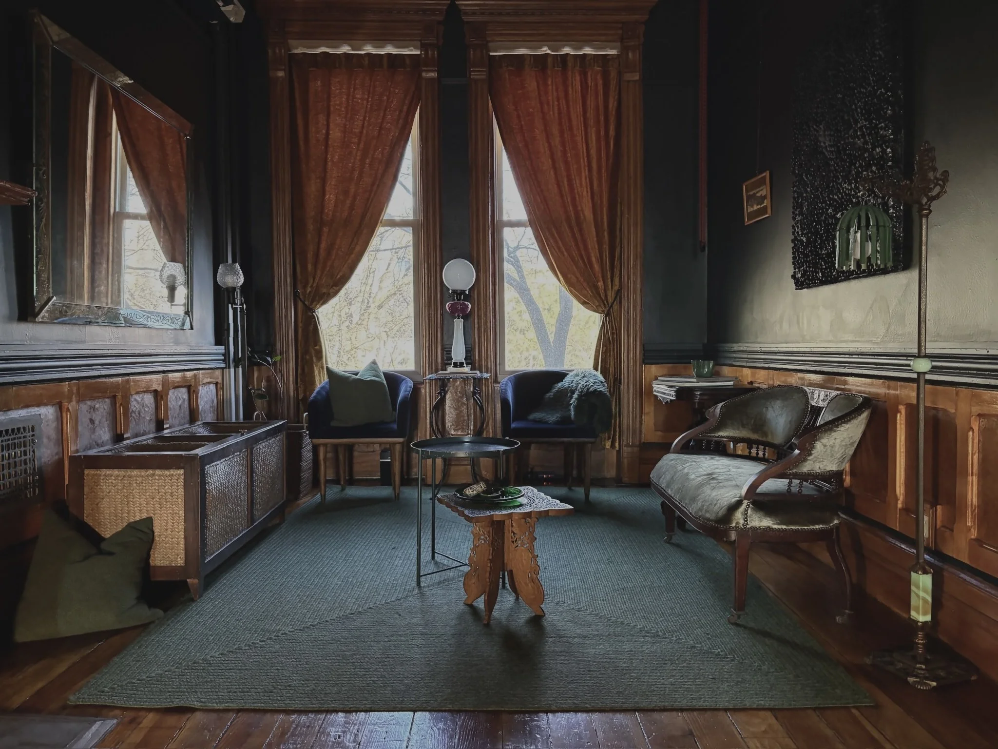 A vintage-style living room with dark wood paneling, large windows with gold curtains, a green rug, and antique furniture including armchairs, a side table, and a floor lamp.