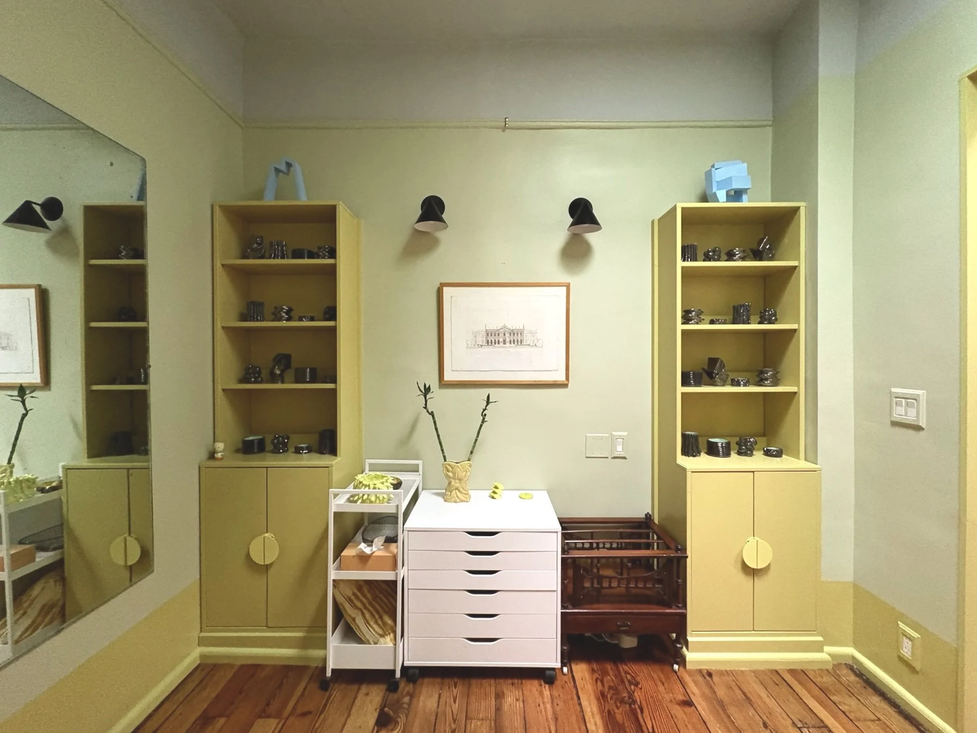 A wall with two yellow bookshelves filled with black decorative objects, a white mobile drawer unit, a wooden cart, and a framed picture in the center. Vases with branches are on top of the drawers.