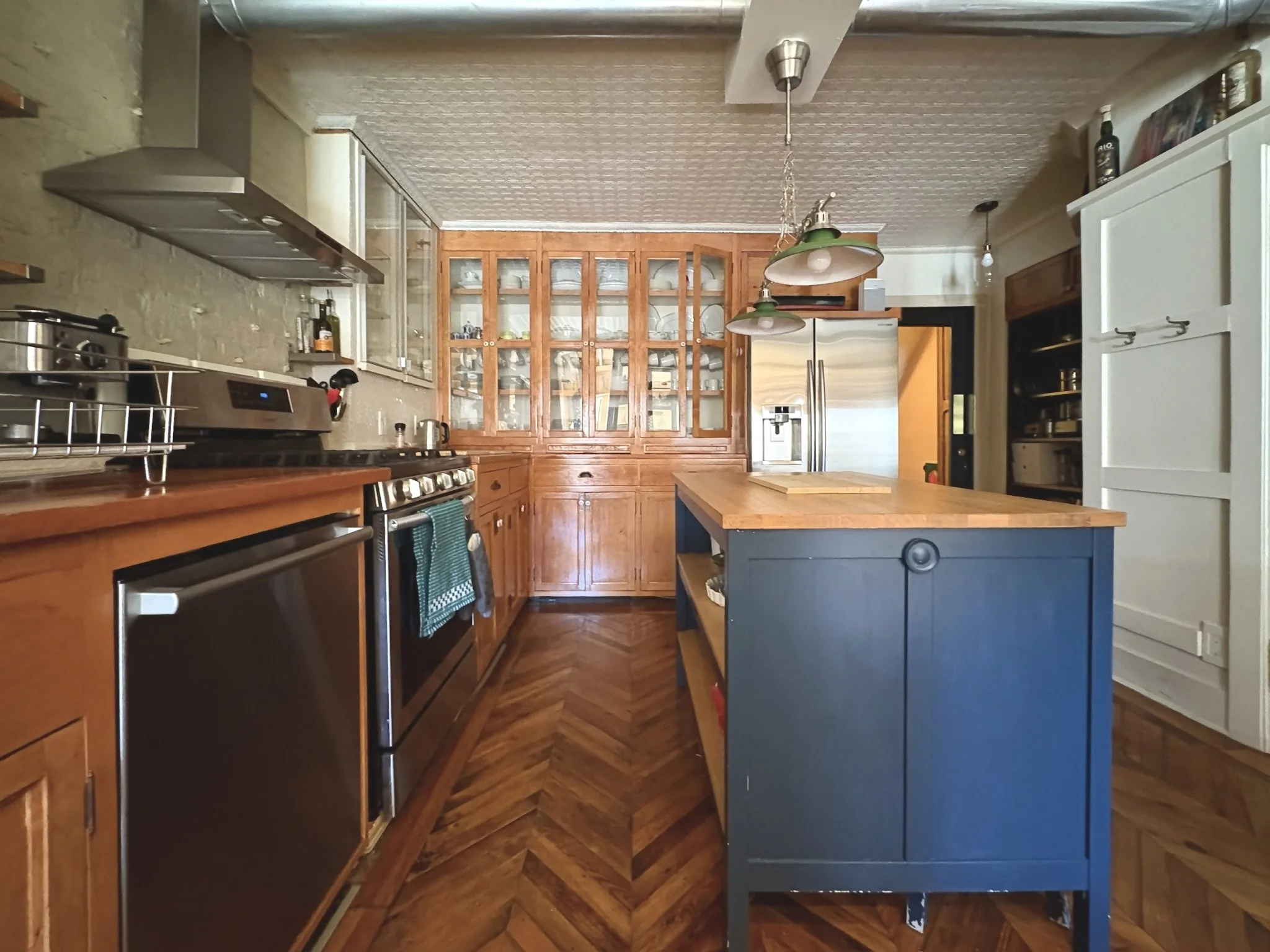 Kitchen with wooden cabinets, stainless steel refrigerator, and a blue island in the center. Wooden flooring and hanging light fixtures are also visible.