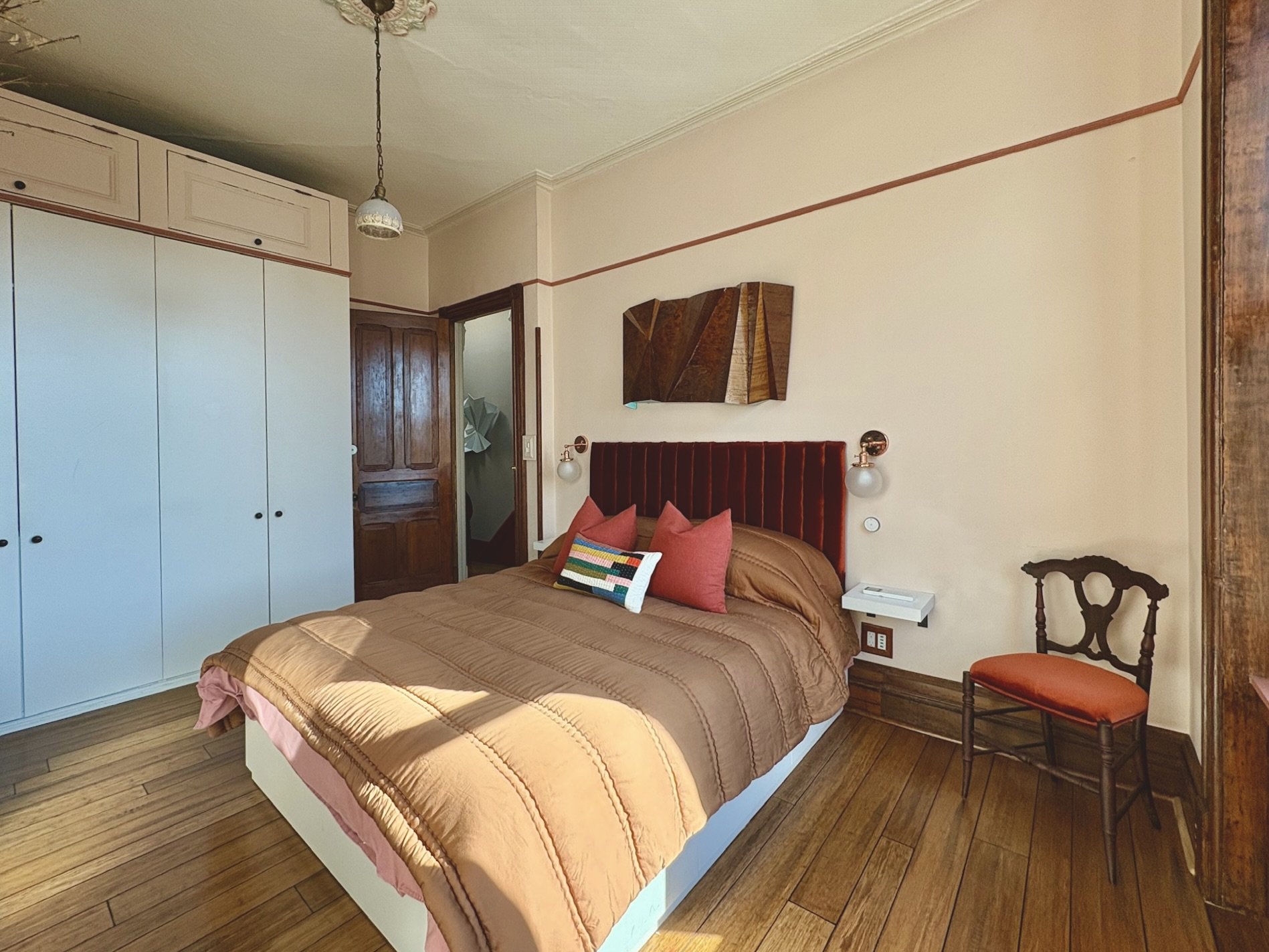 A bedroom with a bed, pillows, and wall decor. Wooden flooring, a white wardrobe, and a brown door. A vintage chair with an orange cushion and a wall-mounted light fixture.