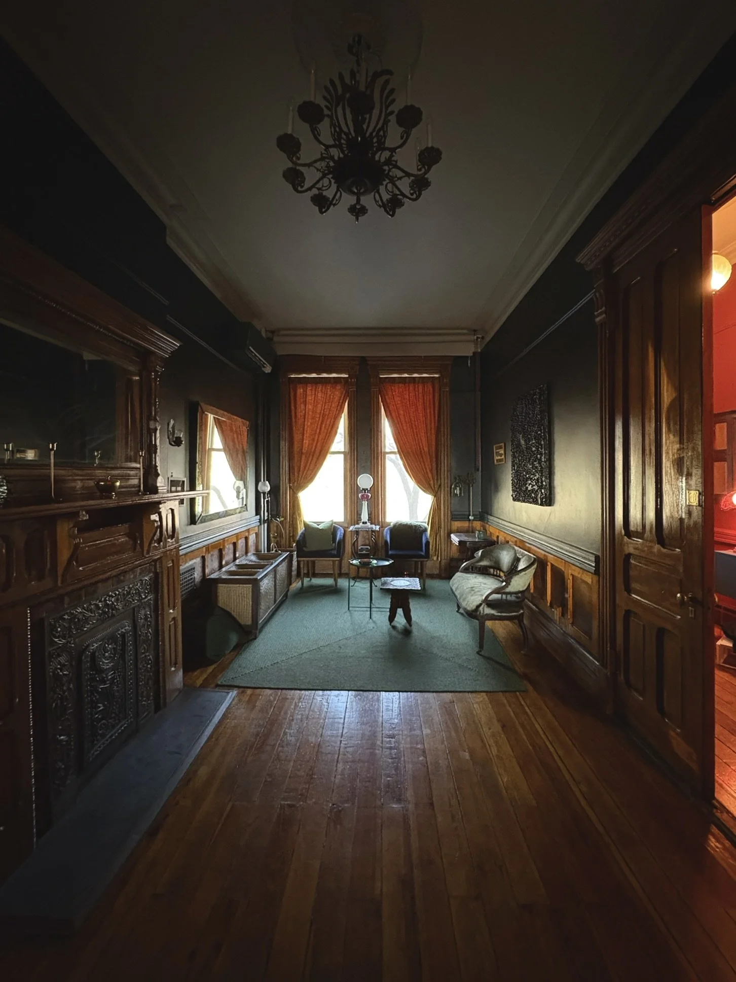 A dark, vintage-style living room with wooden paneling, ornate woodwork, and a chandelier hanging from the ceiling. Large windows with orange curtains let in natural light. The room is furnished with armchairs, side tables, and a green area rug on a 