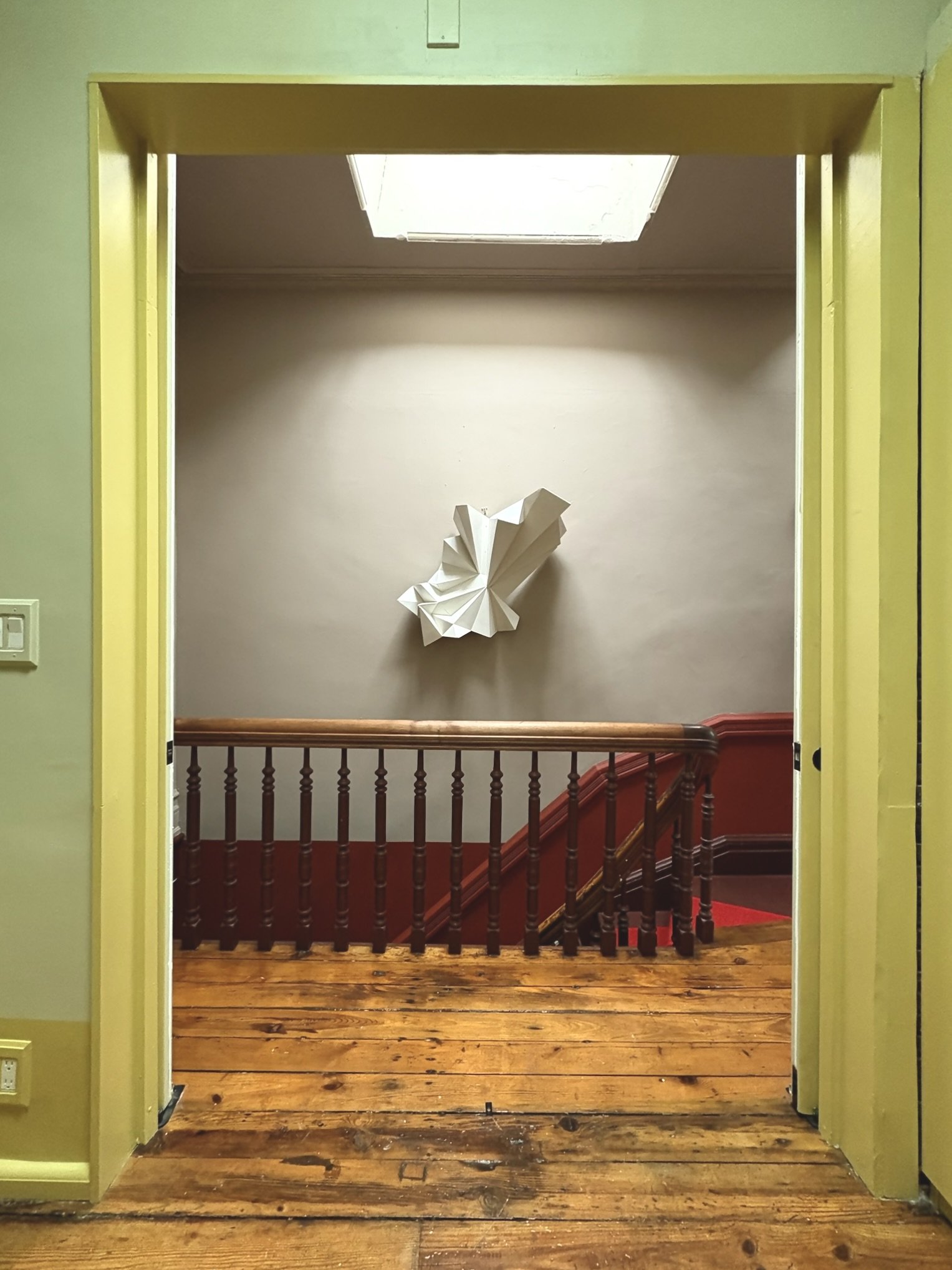 Interior view of a stairway with a wooden railing, a wall art piece resembling a folded paper butterfly, a skylight, and wooden flooring.