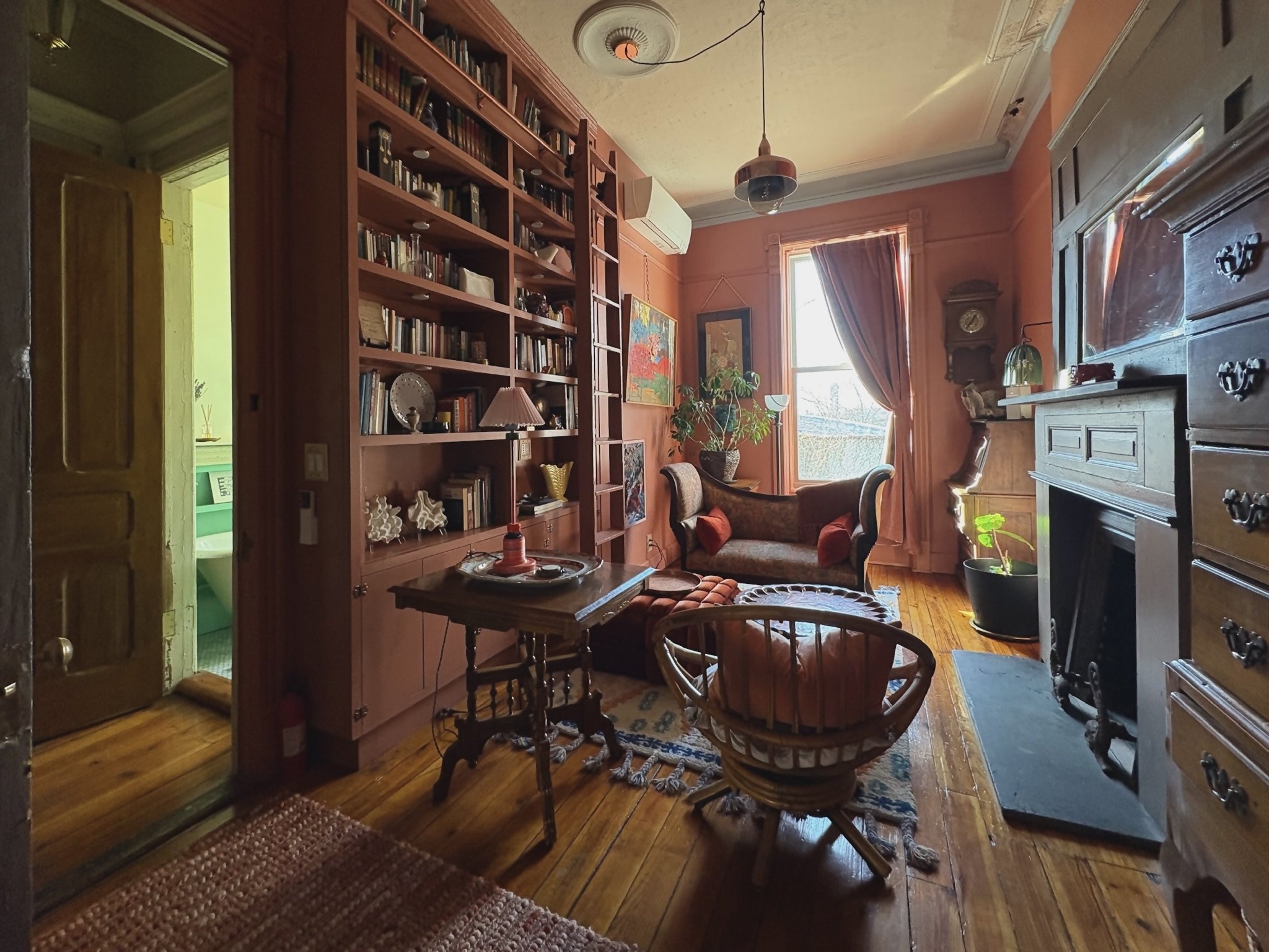 A cozy living room with wooden floors, a fireplace, a large window with curtains, a vintage sofa, a round side table with a lamp, a bookshelf with various items, and plants near the window.