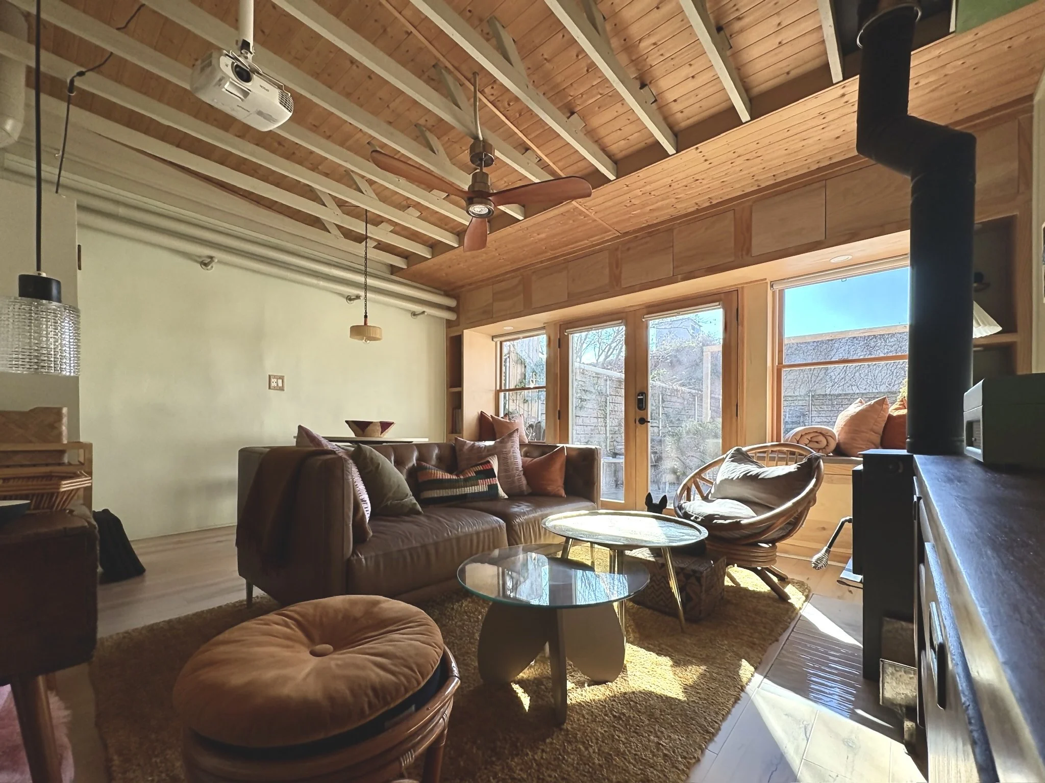 Cozy living room with wooden ceiling, large windows, leather sofa, wicker chair, glass coffee tables, and a round ottoman, illuminated by natural sunlight.