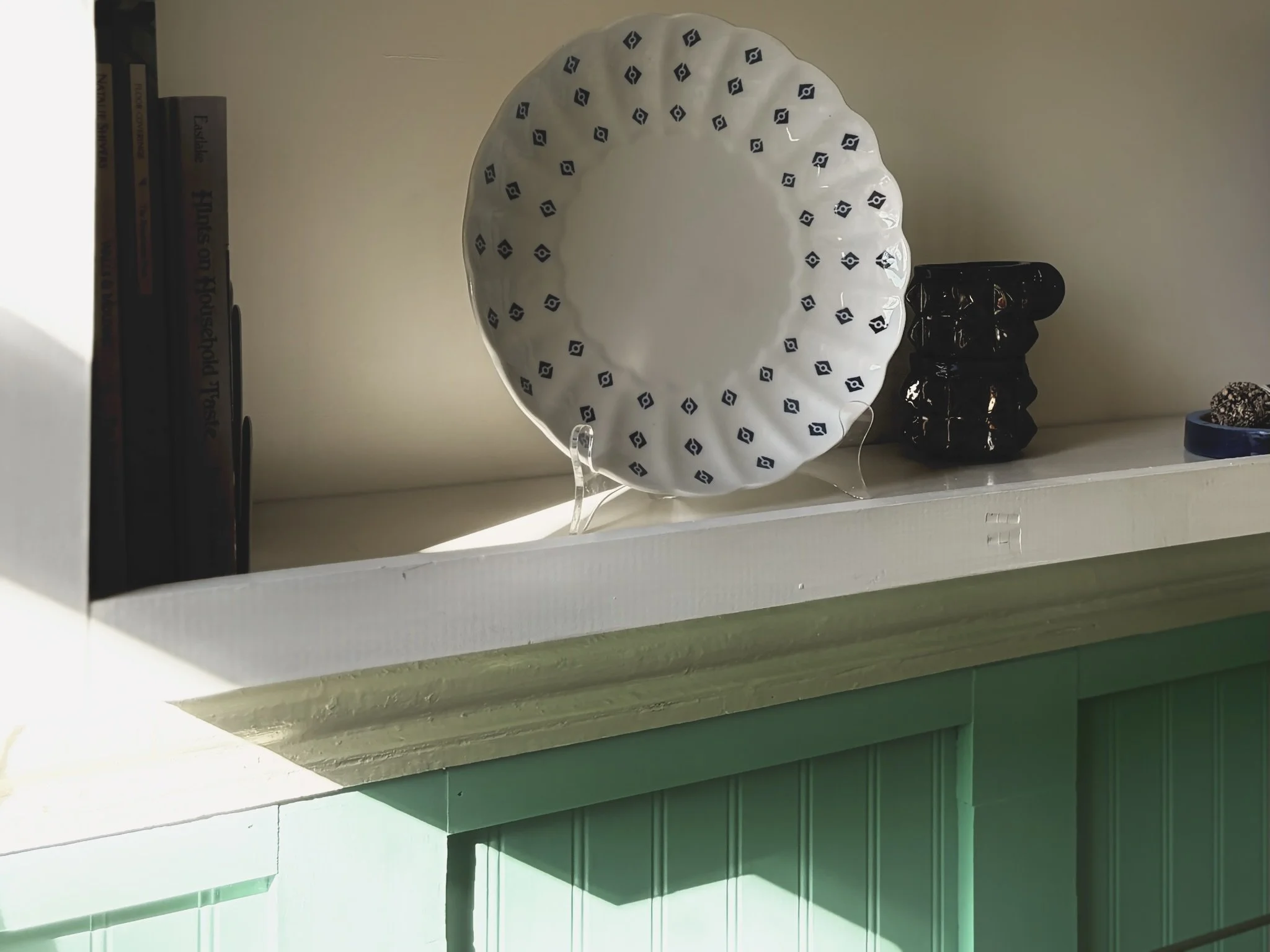 A white decorative plate with a blue geometric pattern around the edge, displayed on a clear stand. To the right, there is a black ceramic sculpture resembling stacked rounded objects. On the far right, part of a small blue dish with a textured object inside. In the background on the left, a bookshelf with books stacked vertically.