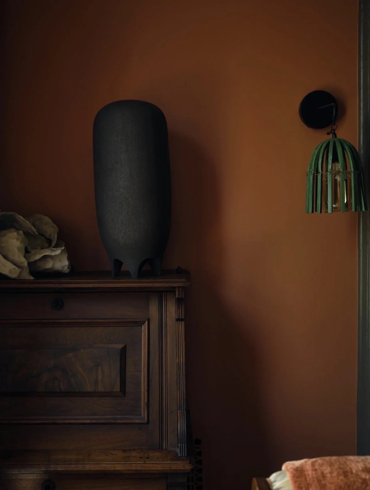 A dark gray sculpture with a rounded top and four small legs is placed on a wooden cabinet against a burnt orange wall. To the left of the sculpture, there is a pile of rocks or stones. On the right side, a black wall sconce with a green and black vi