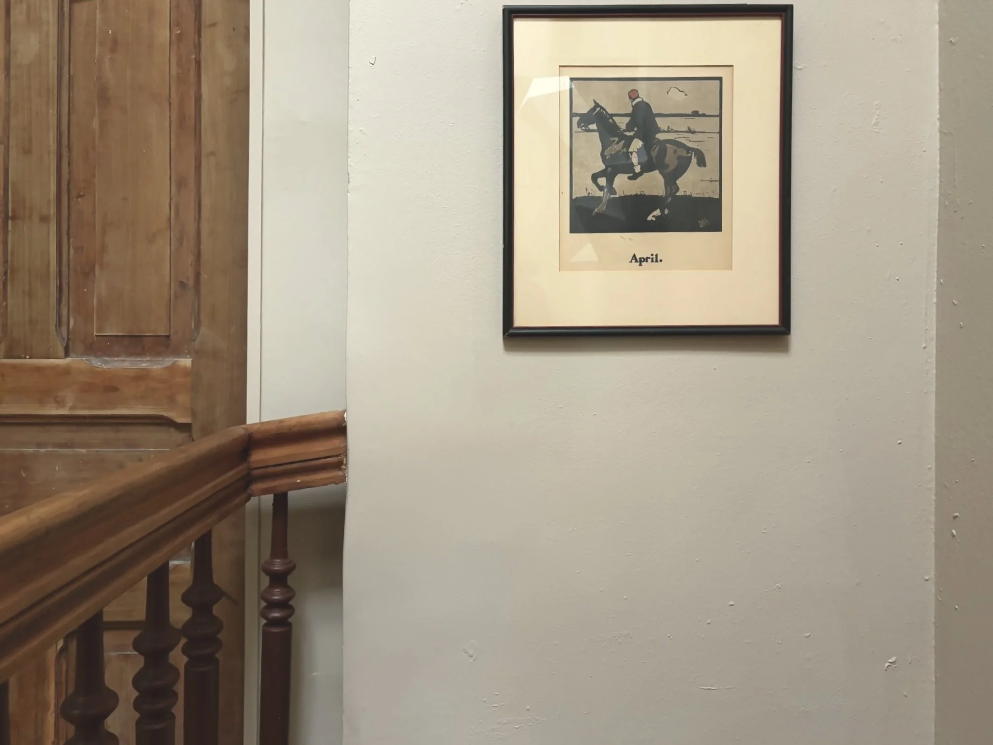 Framed picture of a man riding a horse at a racetrack, with the word 'April' below, hanging on a beige wall in a home or building interior.