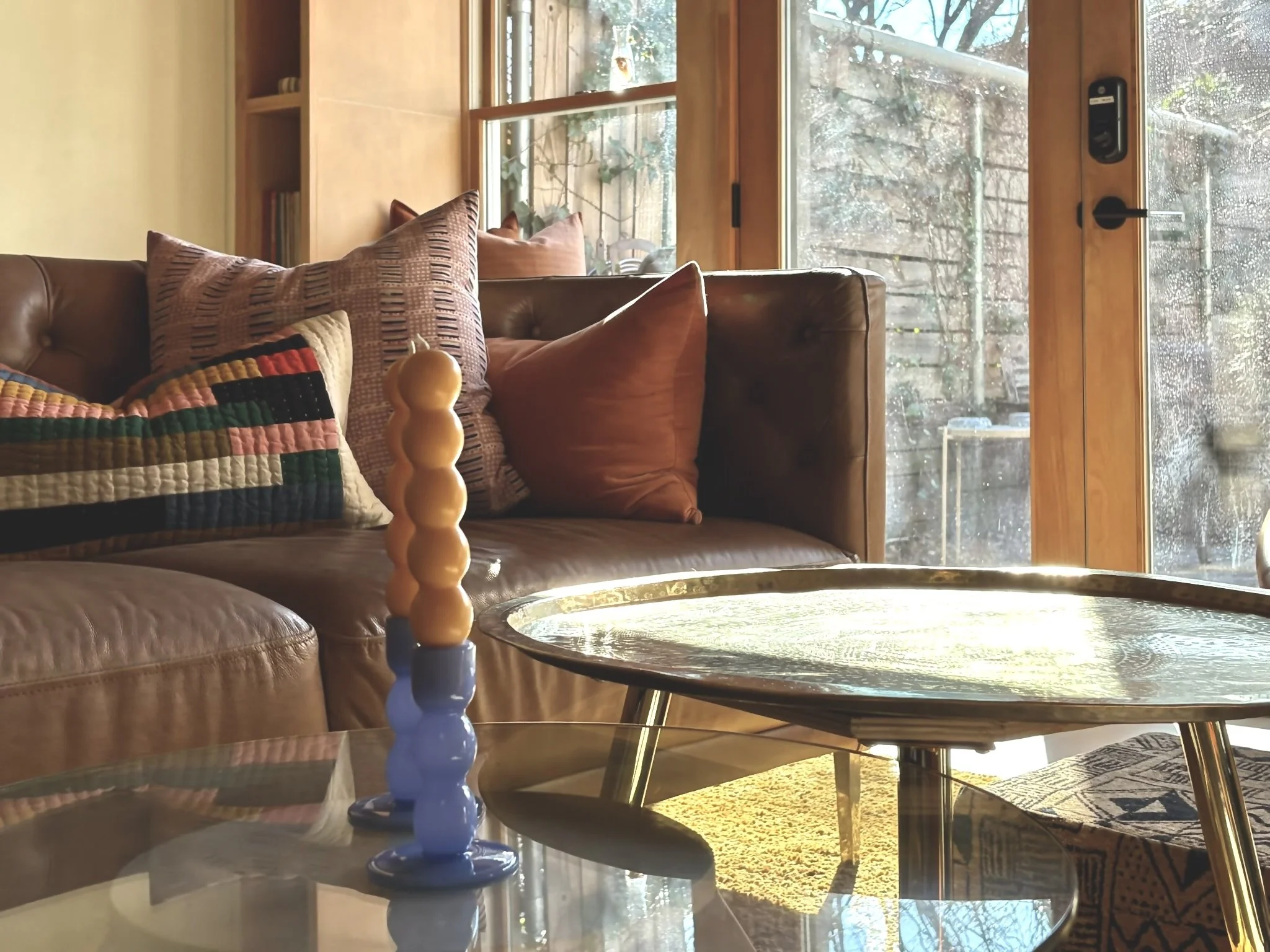 A cozy living room with a leather couch decorated with colorful and textured throw pillows, a glass coffee table with a multicolored decorative candle, and large windows letting in natural sunlight, showing an outdoor patio and garden area.