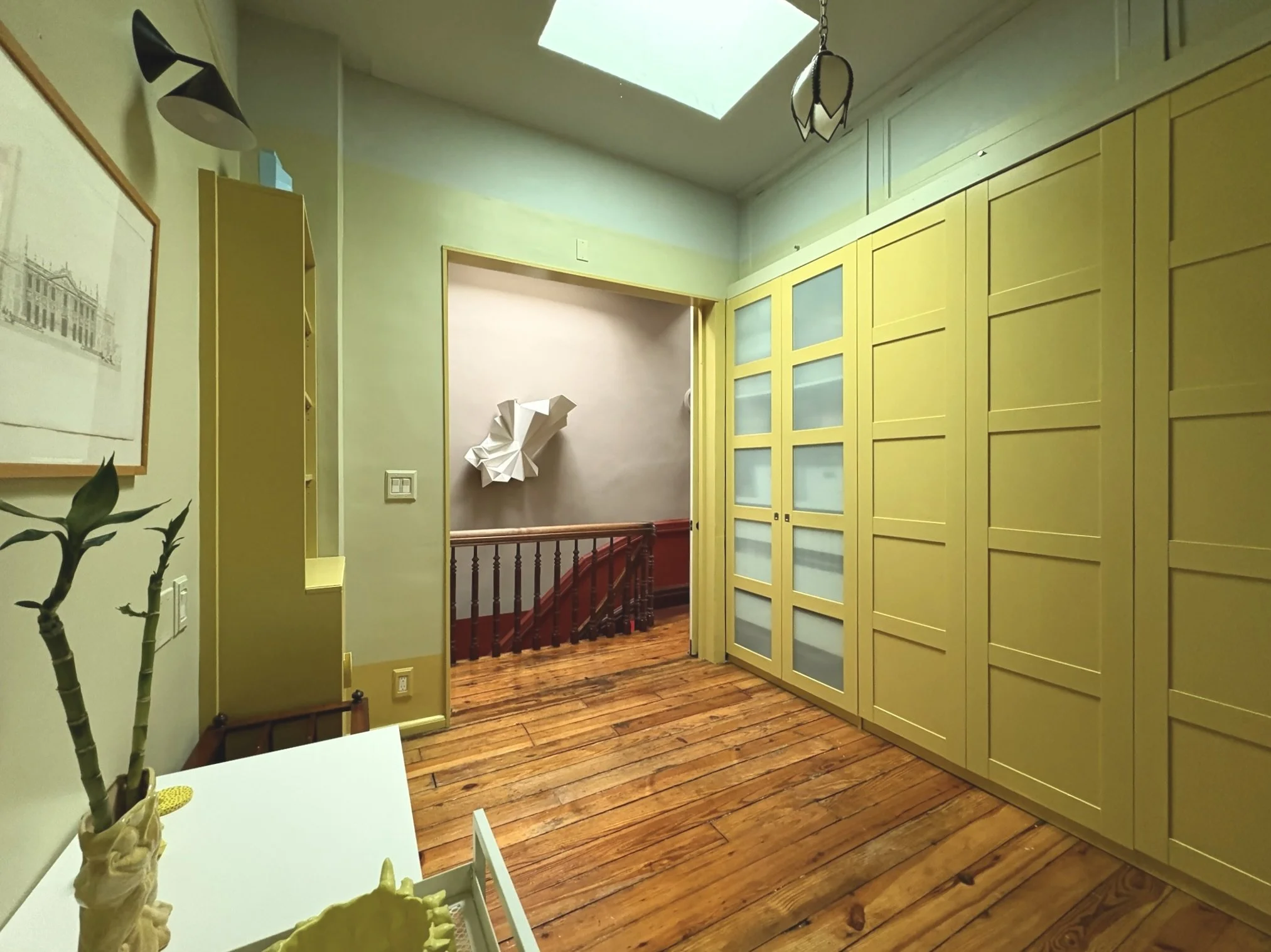 A hallway with yellow cabinets on the right, wooden floor, a small table with a plant on the left, artwork and a paper sculpture on the wall, and a staircase visible through an open doorway.