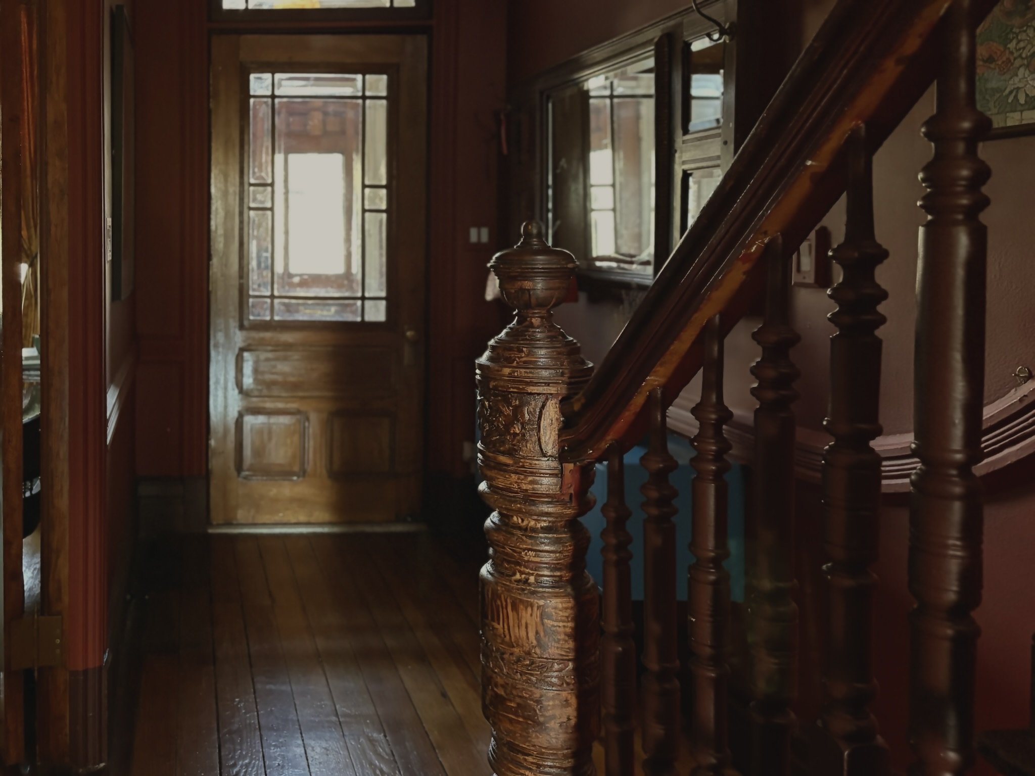 Interior view of a house showing a wooden staircase with turned balusters, a wooden door with a glass window, and wood-paneled walls and flooring.