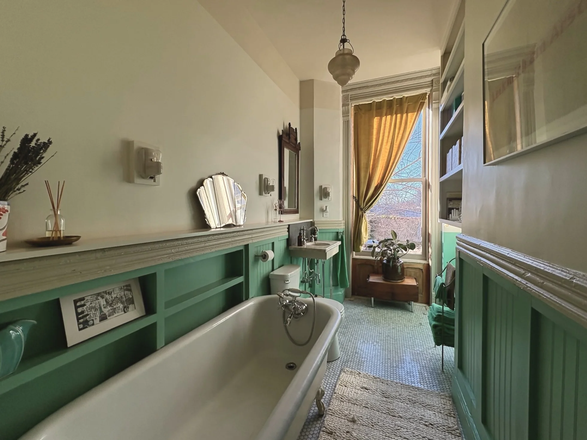 Bathroom with vintage teal wainscoting, clawfoot tub, small sink, window with yellow curtain, potted plant, and patterned floor tiles.