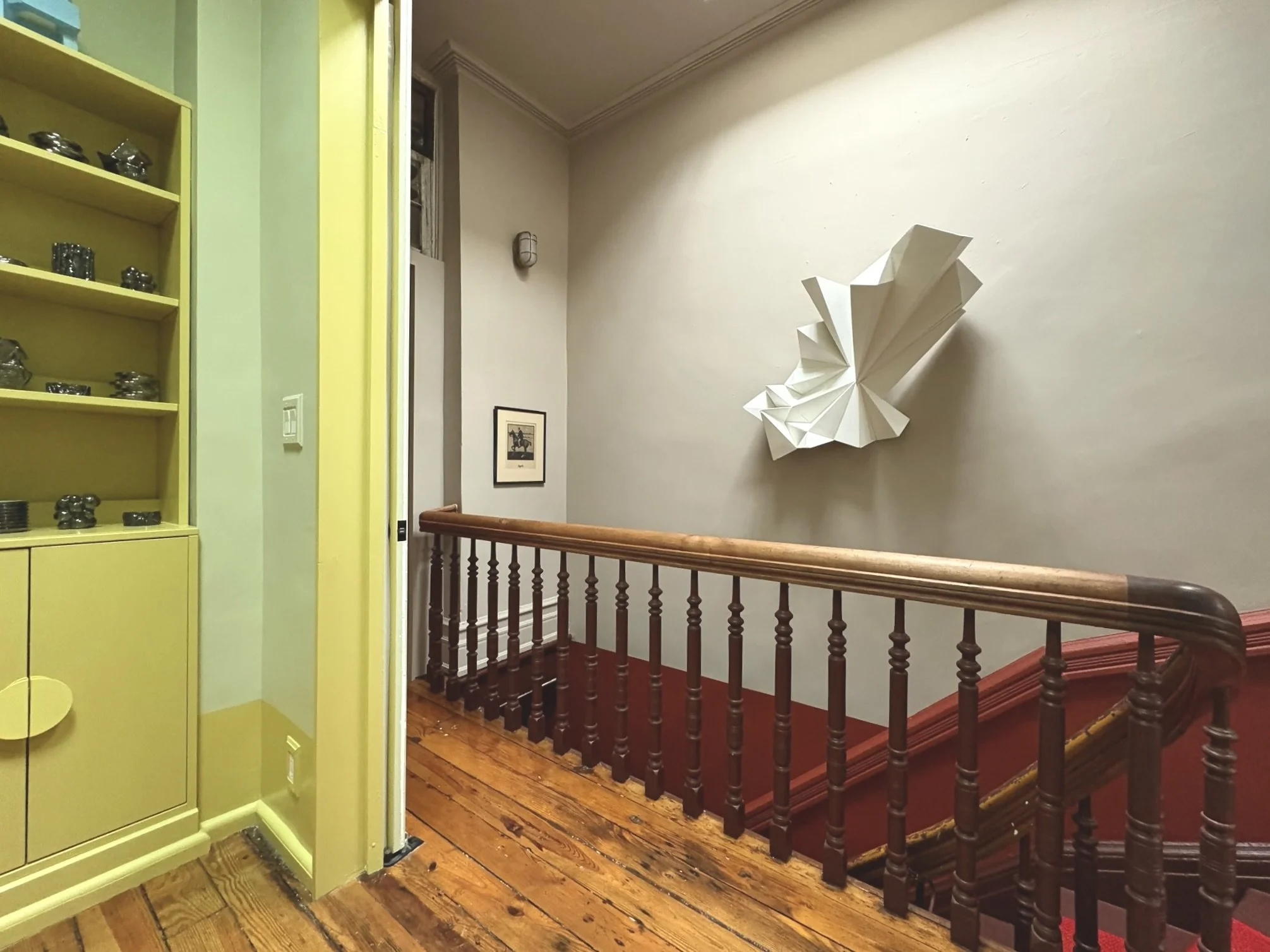 Interior view of a staircase landing with a wooden handrail, a decorative butterfly wall sculpture, framed artwork, and a yellow cabinet with decorative objects.