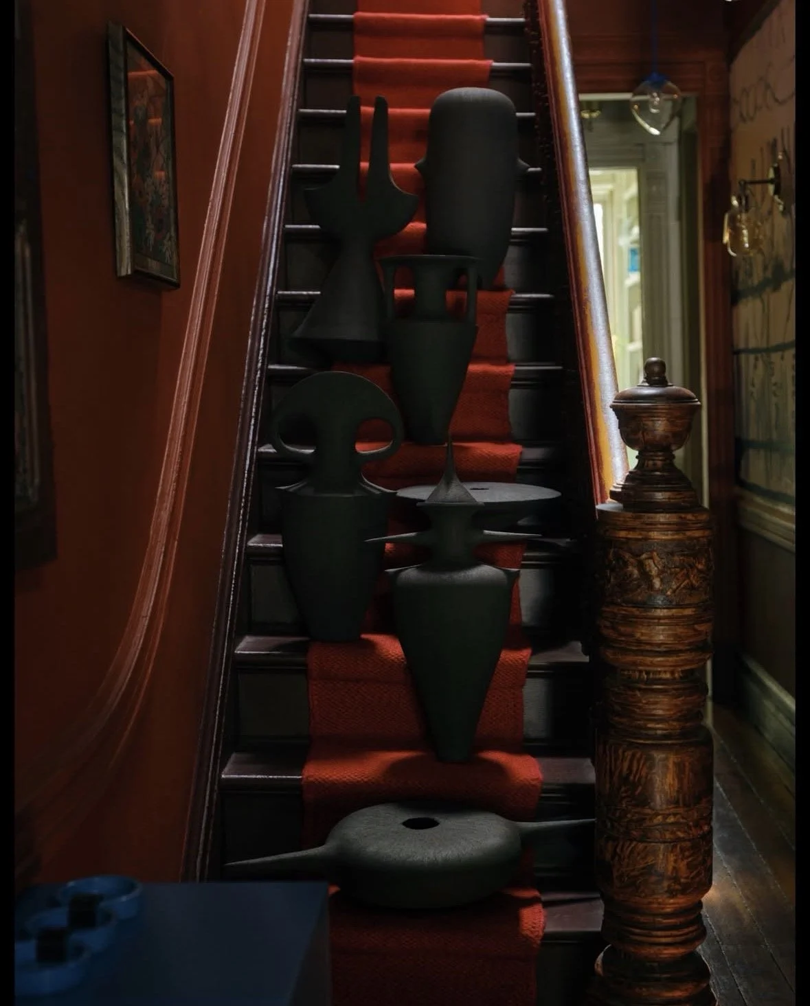 A staircase with a red carpet runner, decorated with several black abstract sculptures placed on the stairs. The staircase has wooden handrails, and the surrounding interior features dark wood paneling and artwork on the walls.