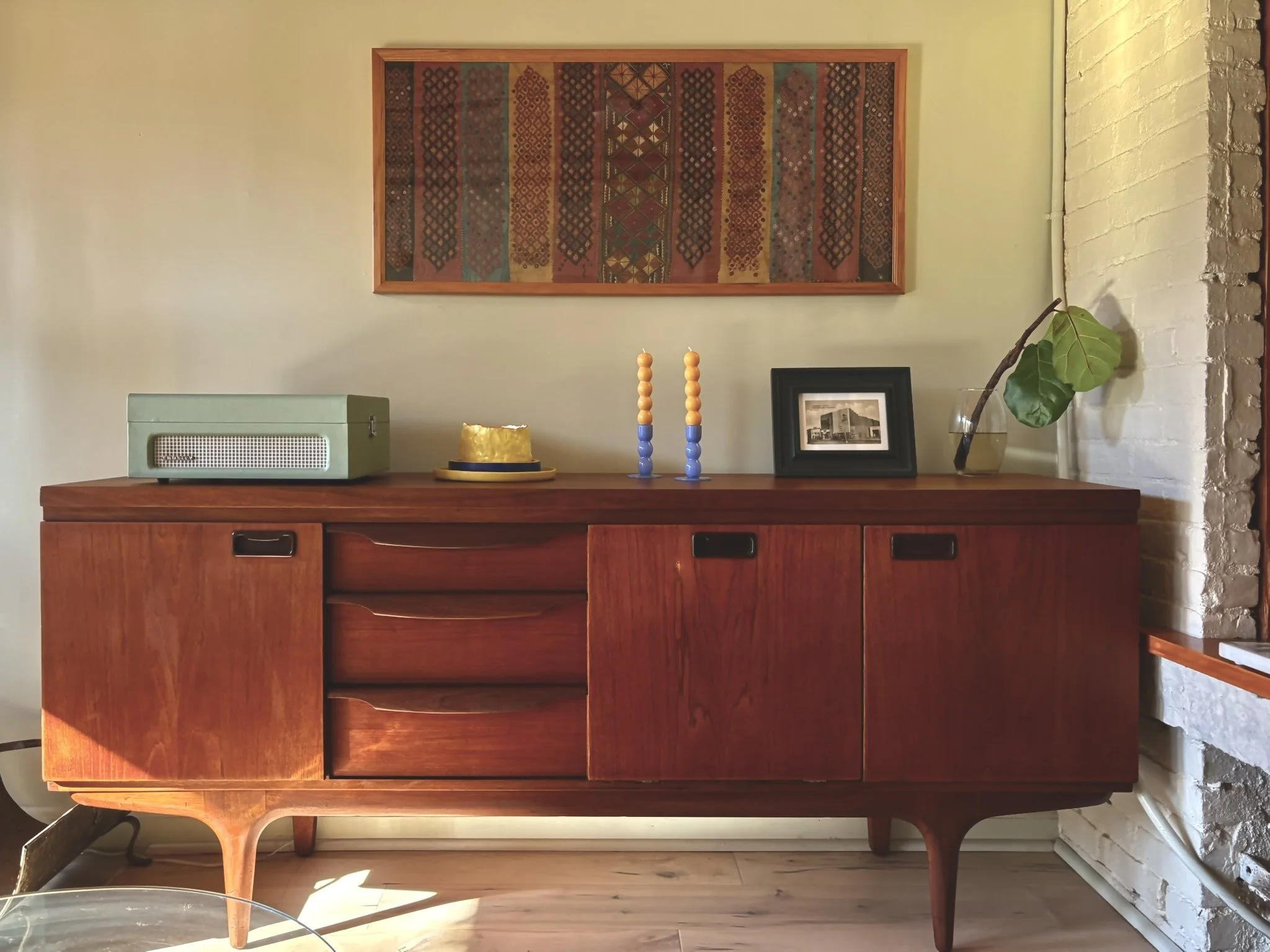 Mid-century wooden sideboard with decorative objects on top, including a green vintage radio, a gold cake, two colored candlesticks, a framed black and white photo, and a glass vase with a large green leaf and branch, against a cream wall with a fram