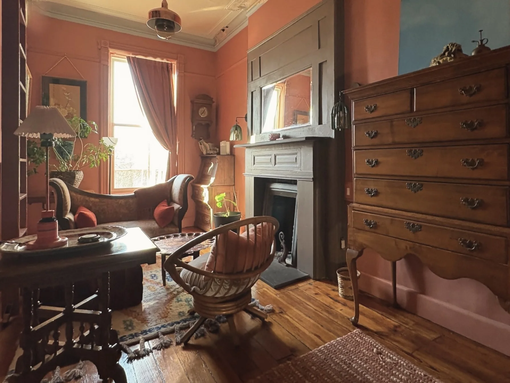 A cozy vintage living room with pink walls, large window with drapes, wooden furniture, a fireplace, and various decor items.