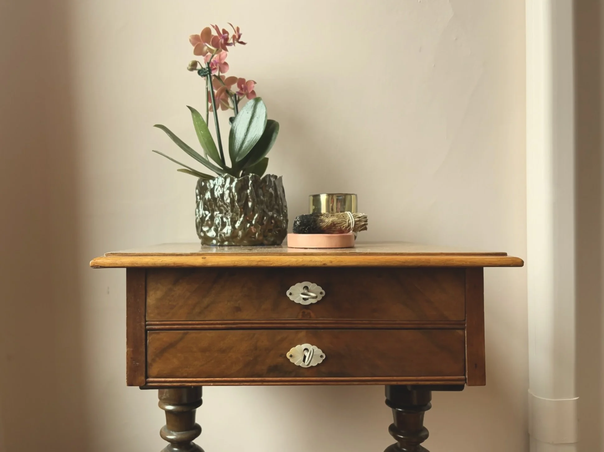 Wooden side table with brass drawer handles, decorated with a potted pink orchid, a small gold container, and a bundle of dried sage.