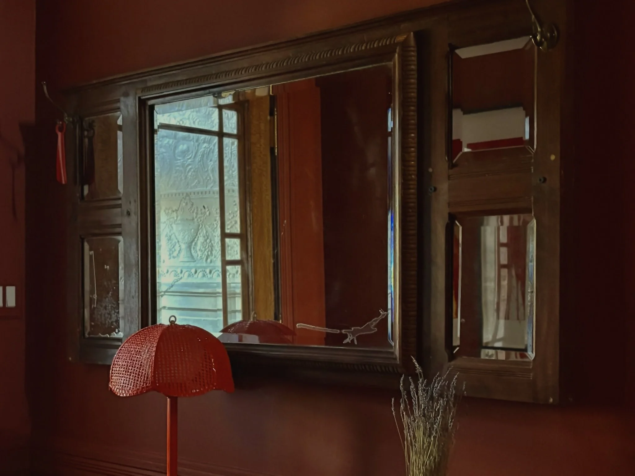 A large antique wooden mirror on a red wall, with small mirrors on each side, reflected window with frosted glass, red table lamp, and dried flowers.