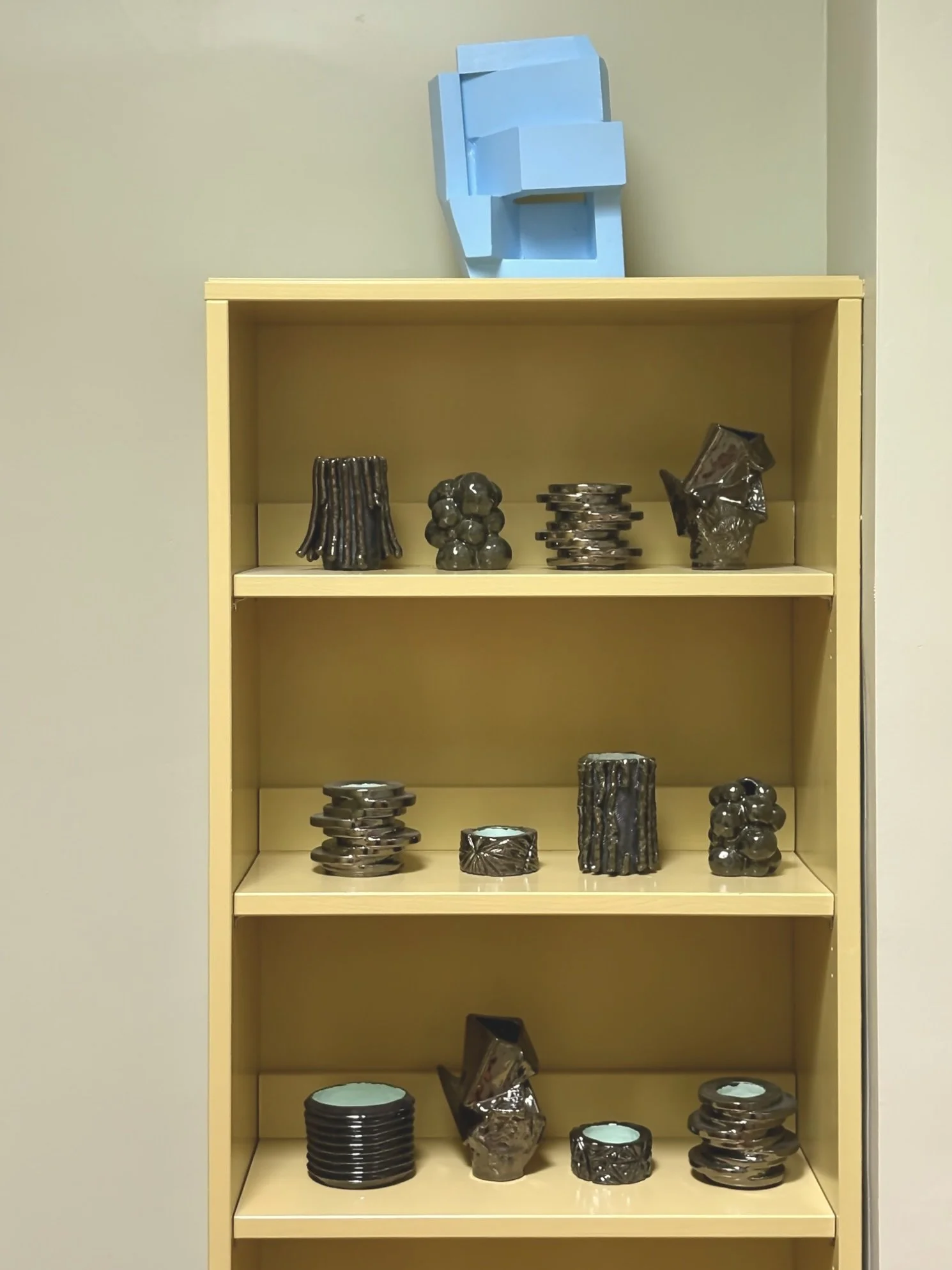 Yellow bookshelf with black ceramic candle holders and decorative objects on three shelves, topped with a blue geometric sculpture.