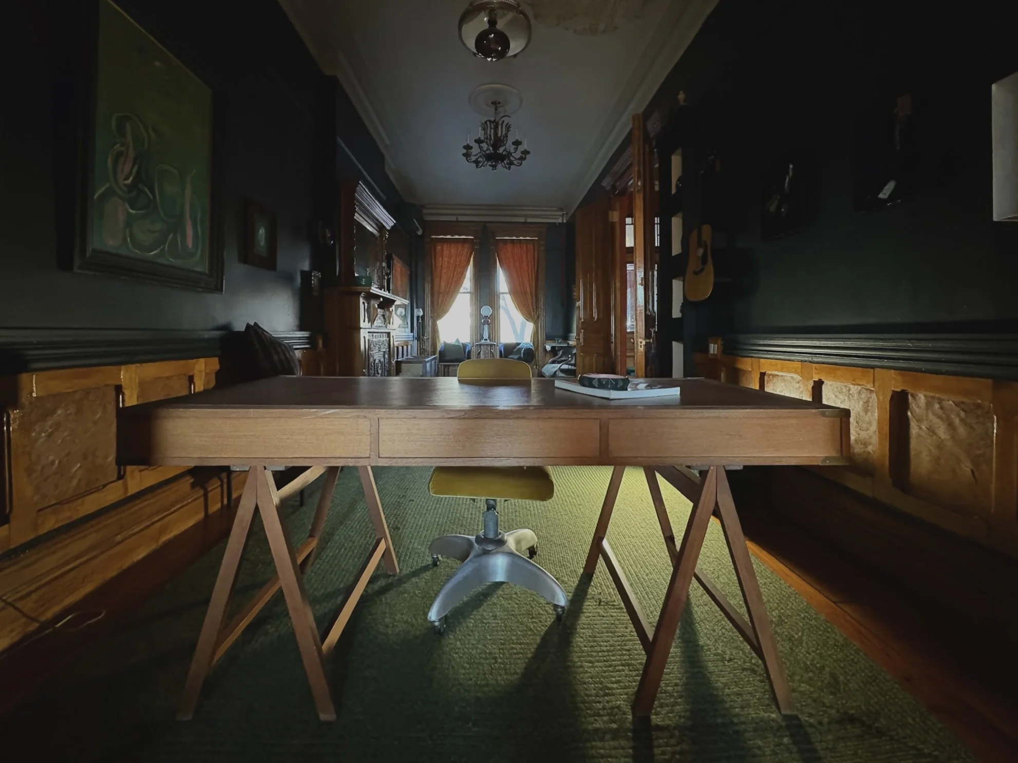 Interior of a room with a wooden table and a yellow office chair, dark walls with artwork, and a window with orange curtains at the far end.