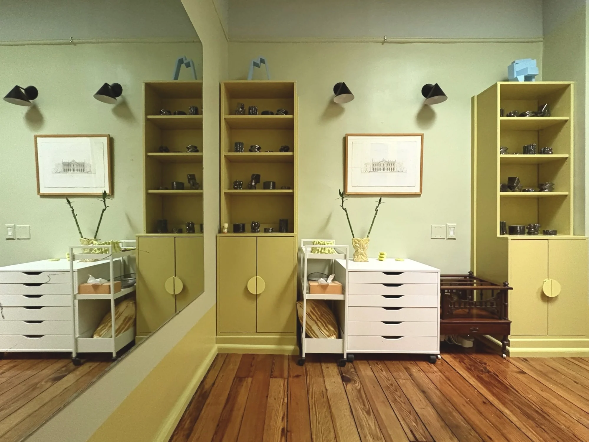 Interior of a room with yellow shelves, black spotlights above, framed artwork on light green walls, and three white and one dark wooden storage units with decorative vases and art pieces.