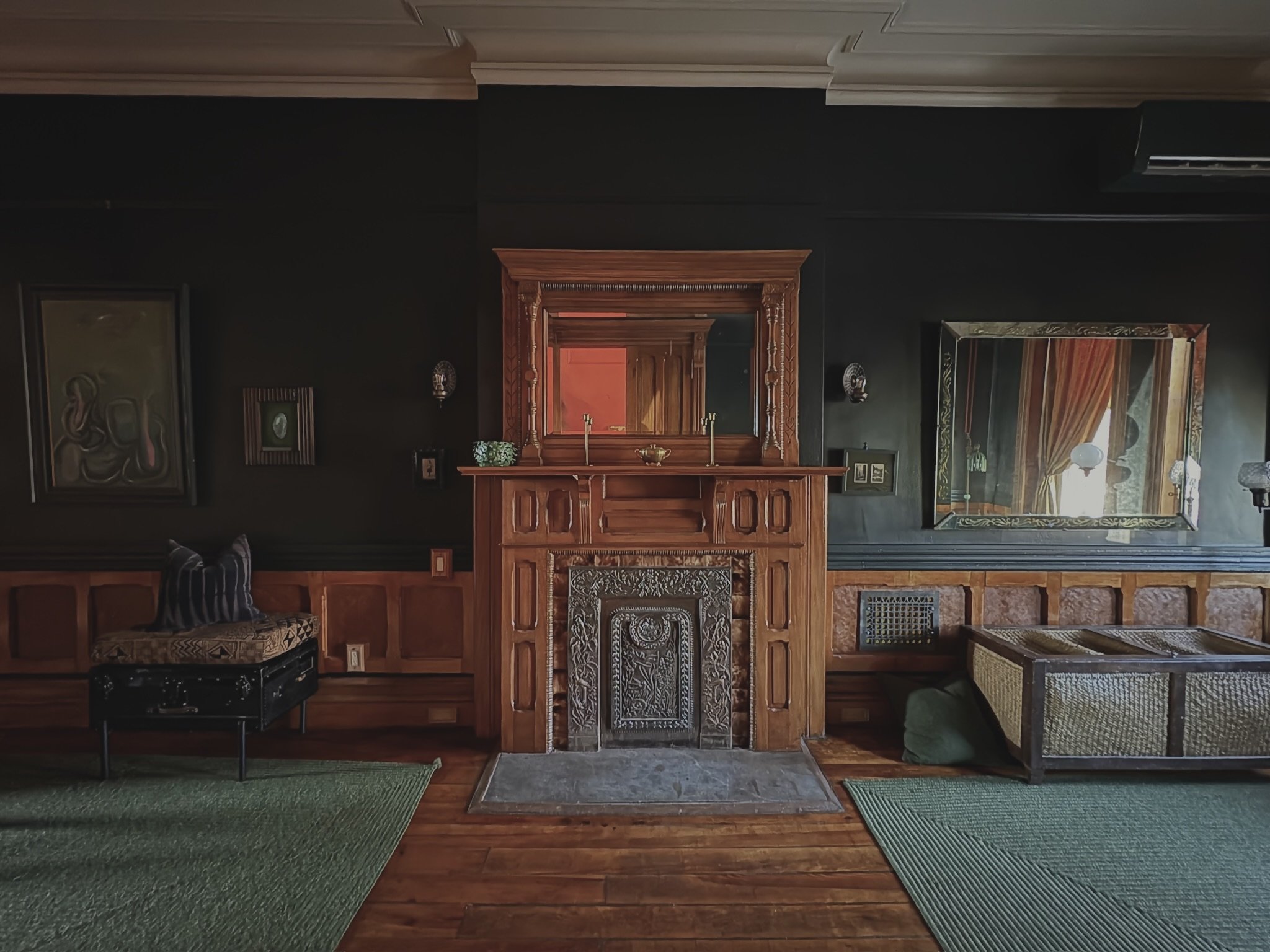 Interior of a room with a dark wall, wooden fireplace mantel with ornate details, mirrors and paintings on the wall, green rugs on a wooden floor, and a cushioned bench to the right.
