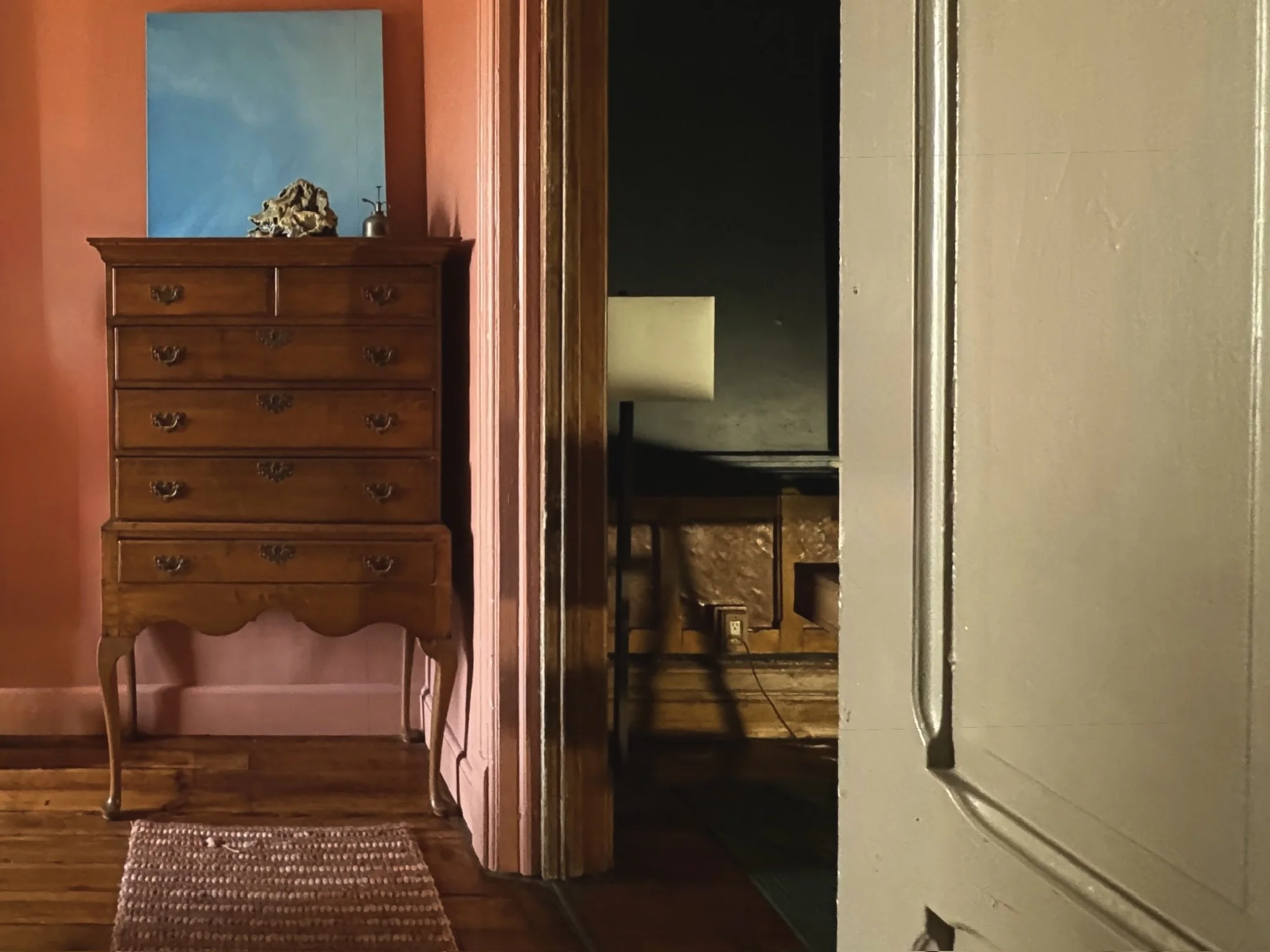 Interior of a room with a wooden dresser against a pink wall, next to a blue painting, with a mirror, lamp, and a rug on a wooden floor.