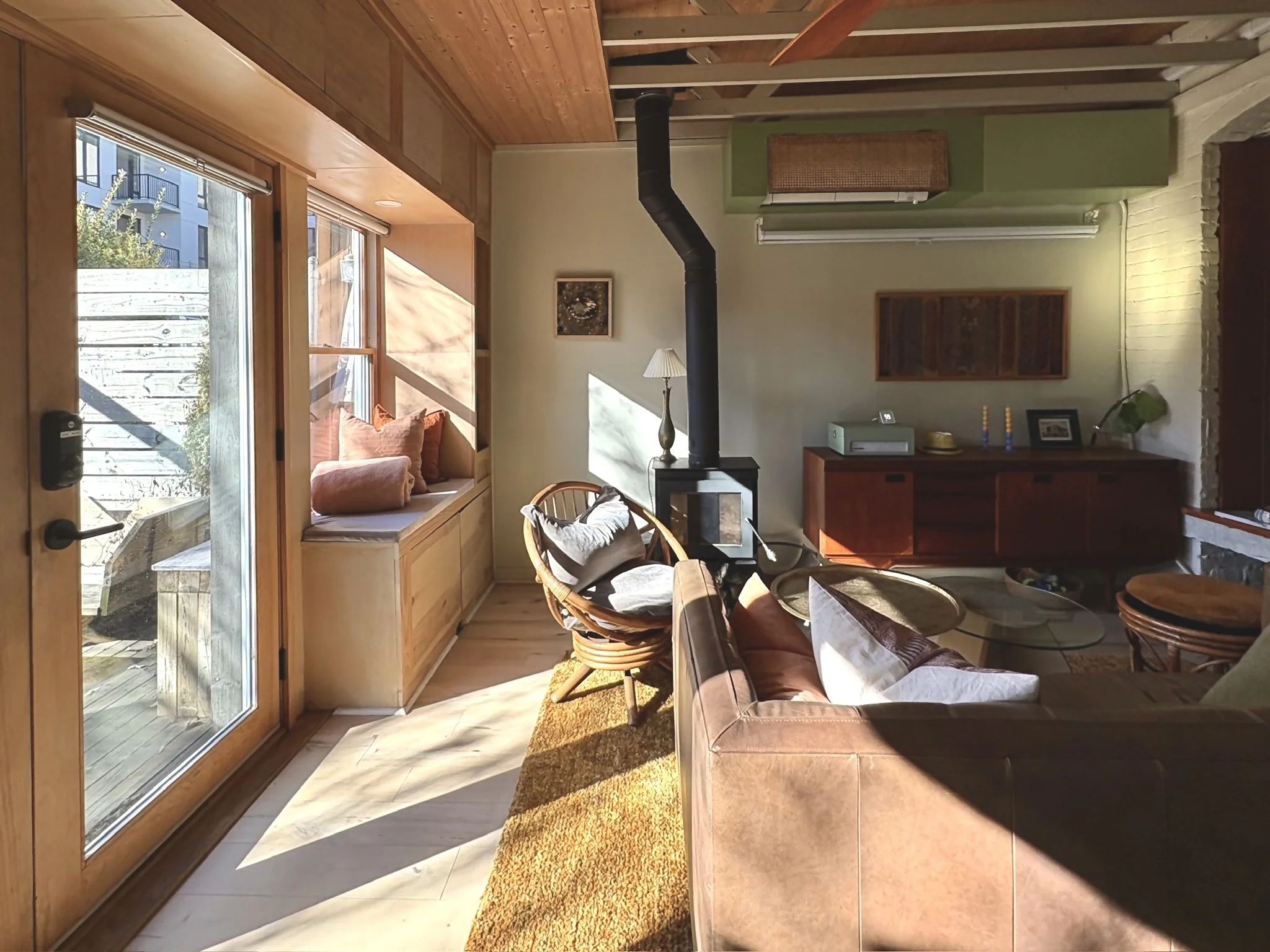 Living room with large glass door, built-in window seat with pillows, wood-burning stove and chimney pipe, wooden furniture, and sunlight.