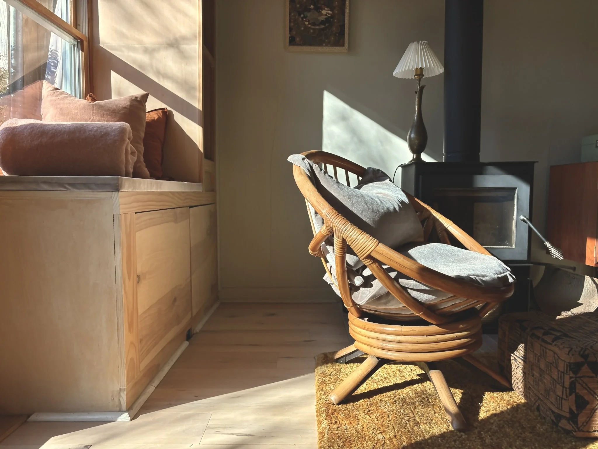 Sunlight streaming through a window illuminating a cozy living room corner with a rattan chair, angled from the side, a dark wood cabinet with a lamp, and a wooden bench with cushions.