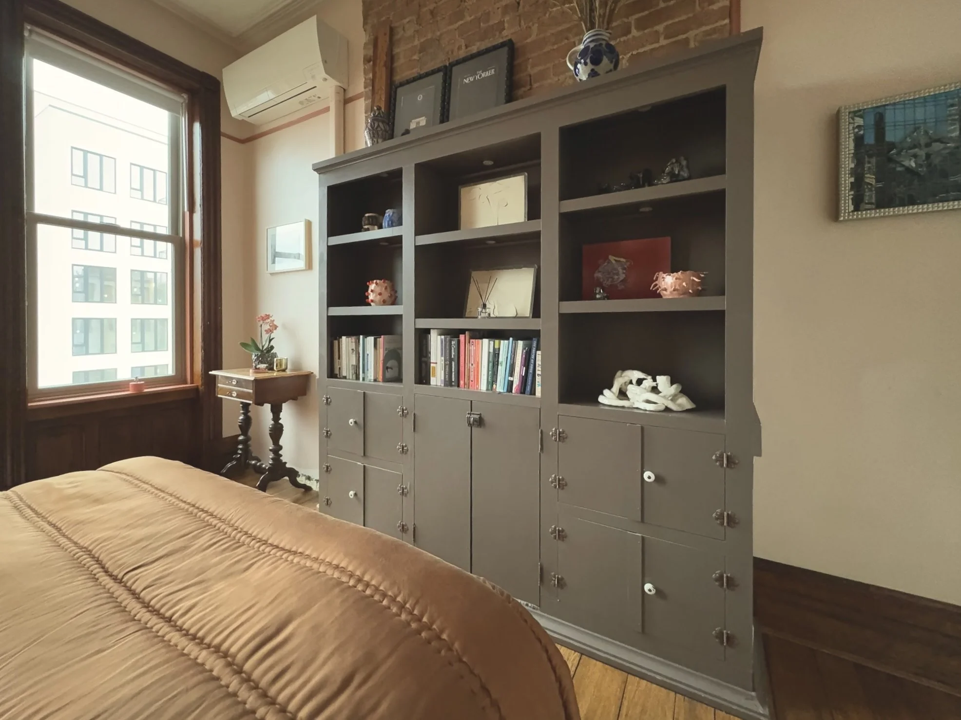 A bedroom with a large gray bookshelf displaying books and decorative items, a window with natural light, a small side table with a potted plant, and part of a bed with a tan or brown comforter.
