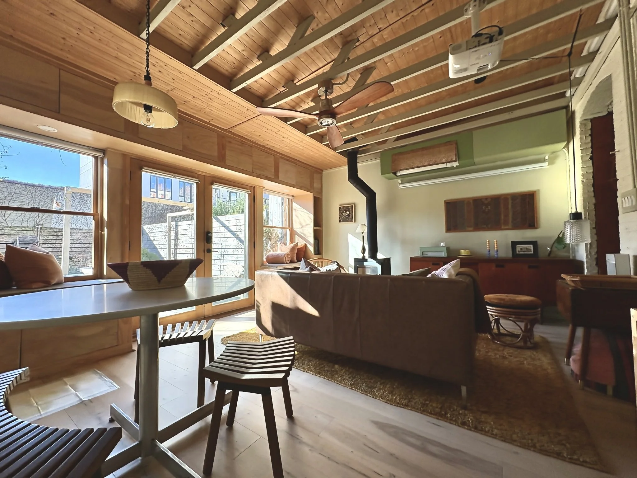 Living room with large windows, wooden ceiling, and furniture, including a brown sofa, armchair, a wood stove, and a sideboard, illuminated by sunlight.