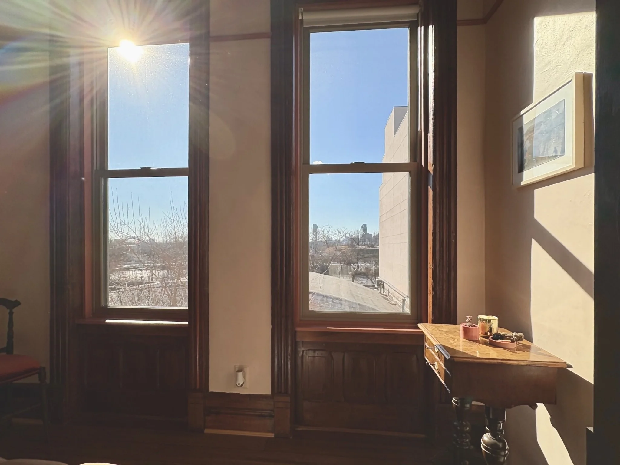 Sunlight streaming through two large windows in a room with wooden trim, yellow walls, a small wooden table with candles and decorative items, and a framed picture on the wall.