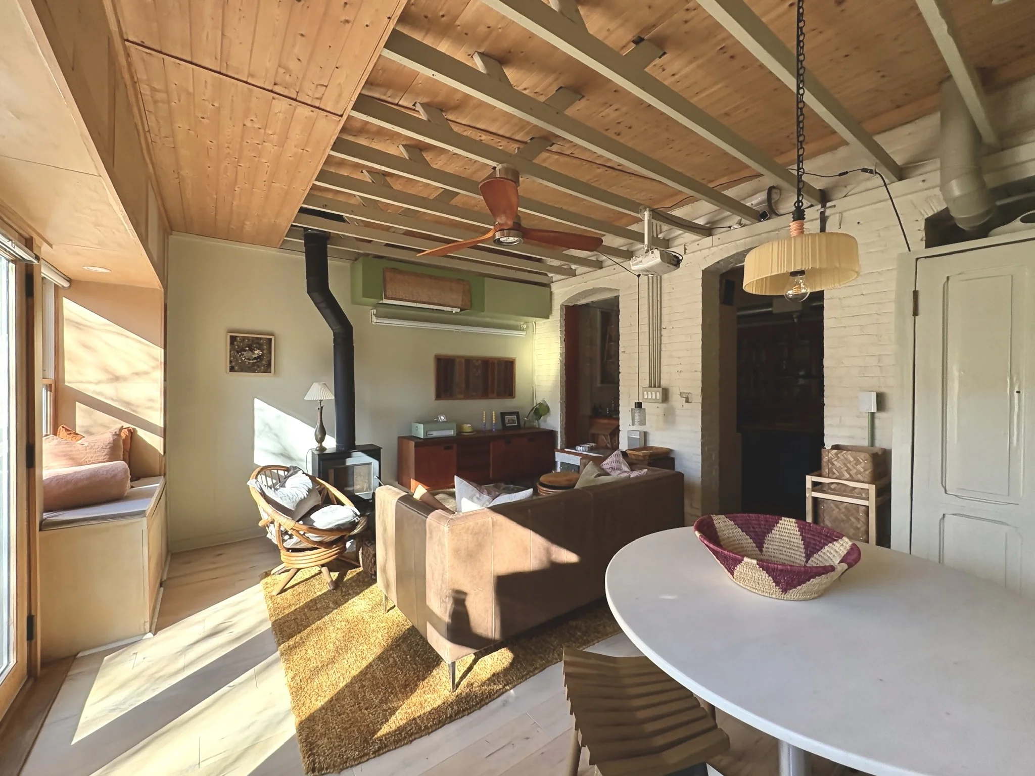 Living room with beige sofa, wooden chairs, and a white coffee table, featuring a wood stove with a black chimney pipe, large windows letting in sunlight, and a ceiling with exposed beams and a ceiling fan.