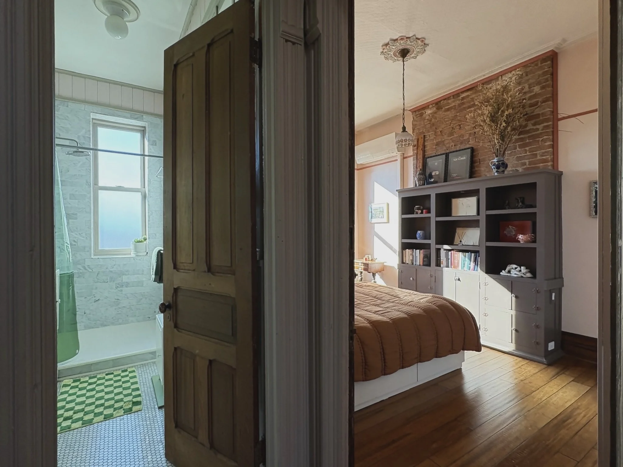 View of a cozy bedroom with a large bookshelf against a brick accent wall, a bed with an orange blanket, a hanging light fixture, and a hardwood floor. The adjacent bathroom is visible through an open door, featuring a frosted window, a shower with m