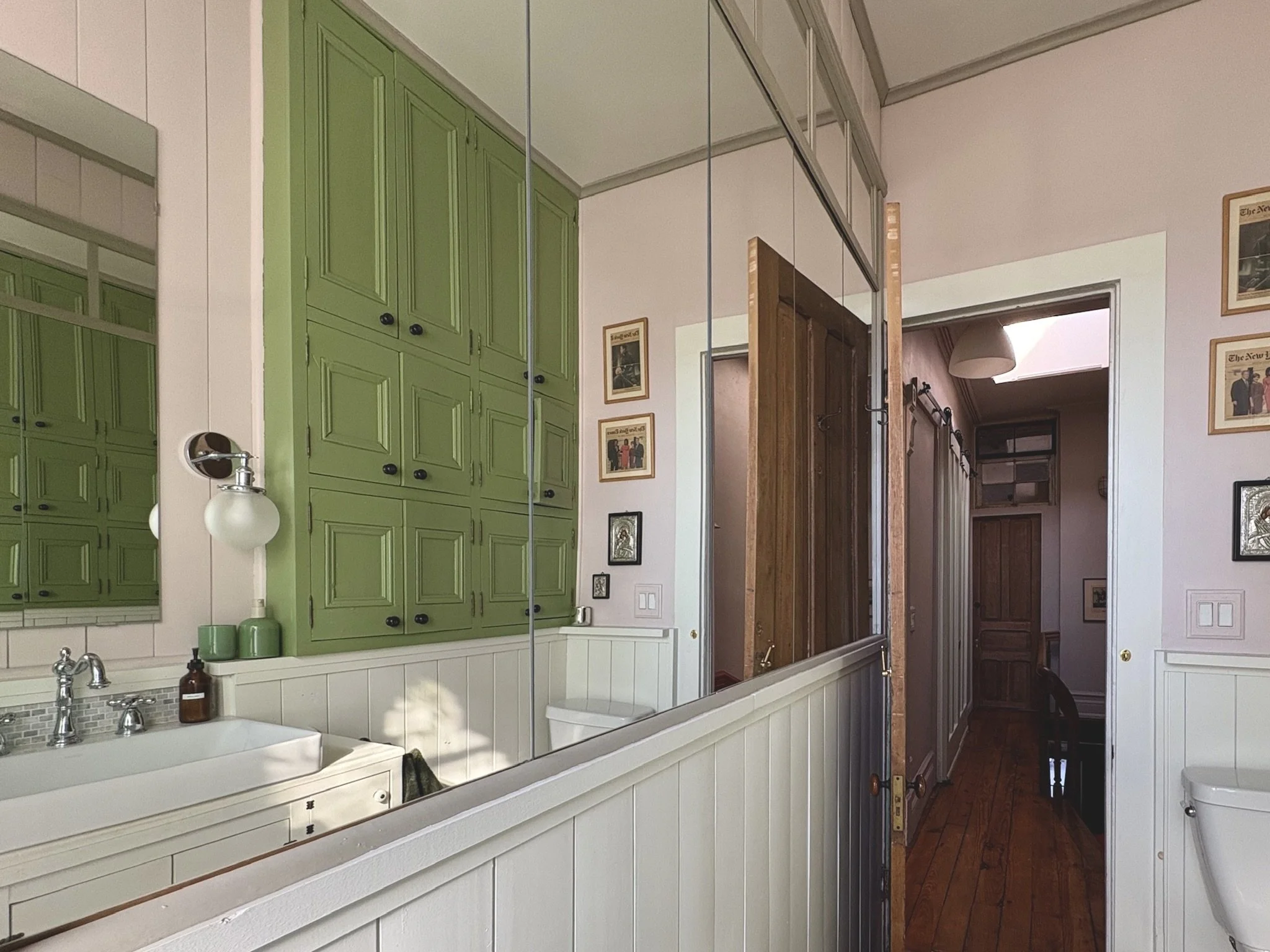 A bathroom with green cabinets, a mirror, a white sink, and framed pictures on the wall, with a wooden door leading to a hallway with wooden floors.