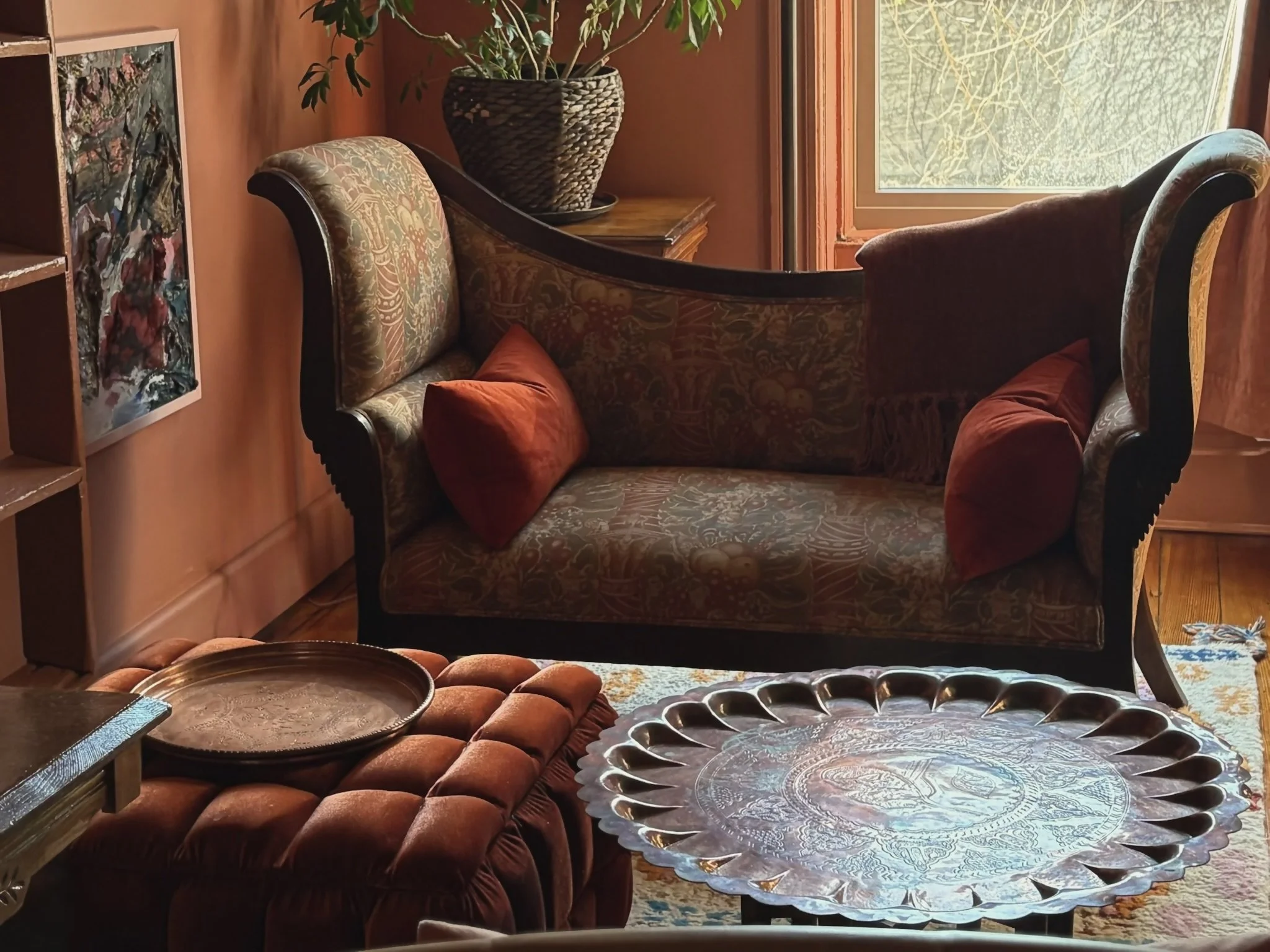 A vintage-style living room with a patterned sofa, two red accent pillows, an ornate metal tray table, a velvet ottoman, a large decorative tray, a wall-mounted painting, a window with a view of green foliage, and a potted plant on a wooden stand.