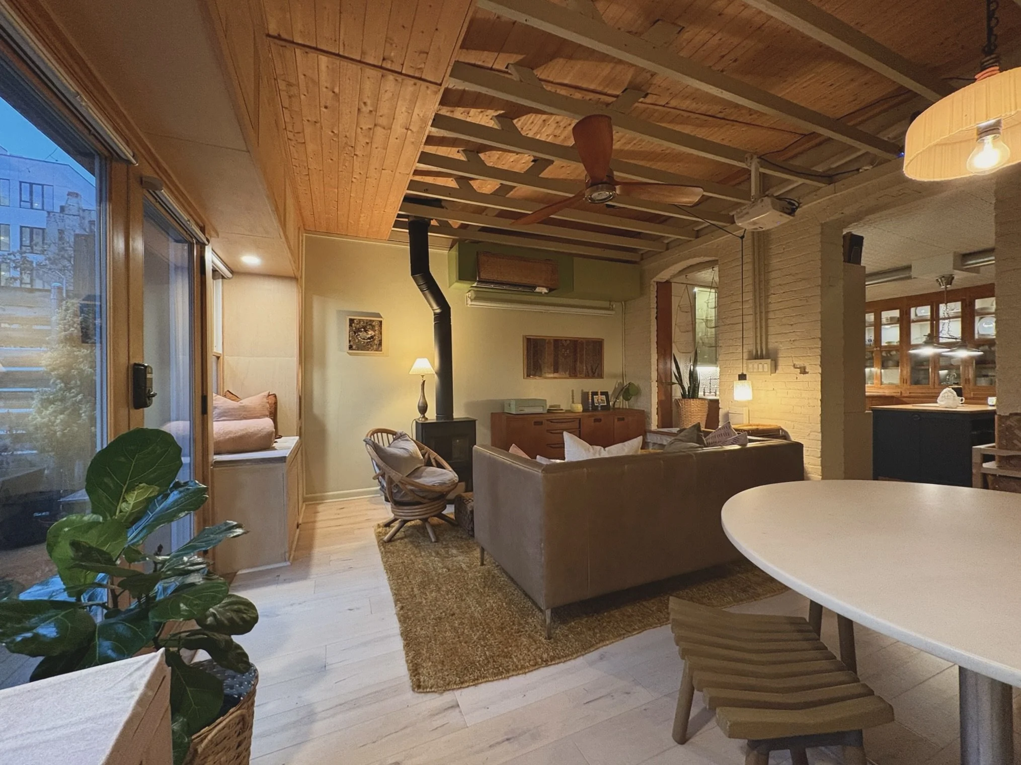 A cozy living room with wooden ceiling and floor, a brown leather sofa with pillows, a wood stove with a black pipe, a small side table with a lamp, a wicker chair, houseplants, a window seat, and a sliding glass door leading outside, illuminated wit