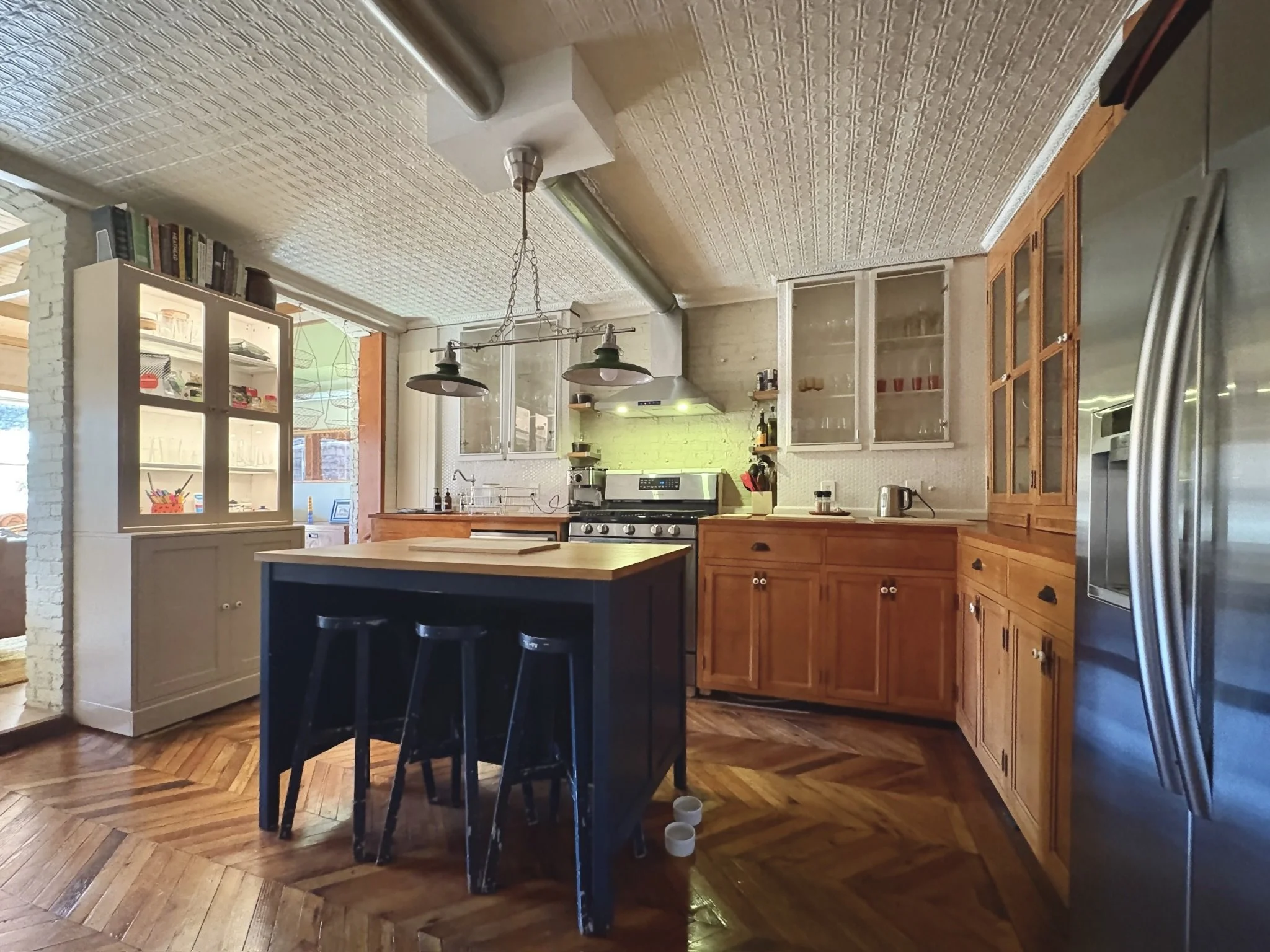 A rustic kitchen with wooden cabinets and a black kitchen island with stools. The kitchen features a stove, chimney hood, and glass-front cabinets, along with various appliances and kitchenware.