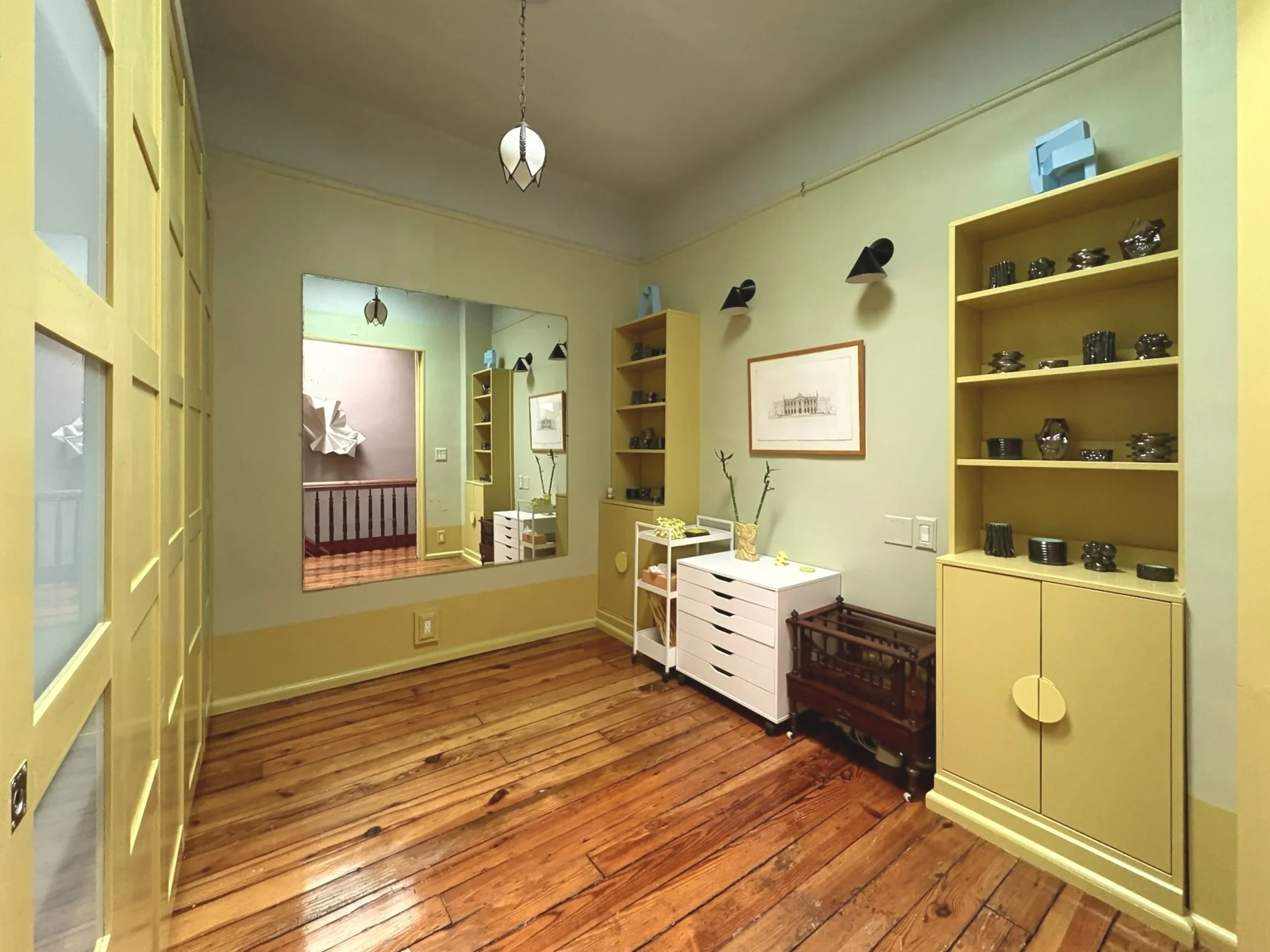 Interior view of a room with light yellow walls and wooden flooring, featuring built-in yellow shelves with black porcelain bowls, a white storage unit, and a framed sketch on the wall, with a large mirror reflecting part of the room.