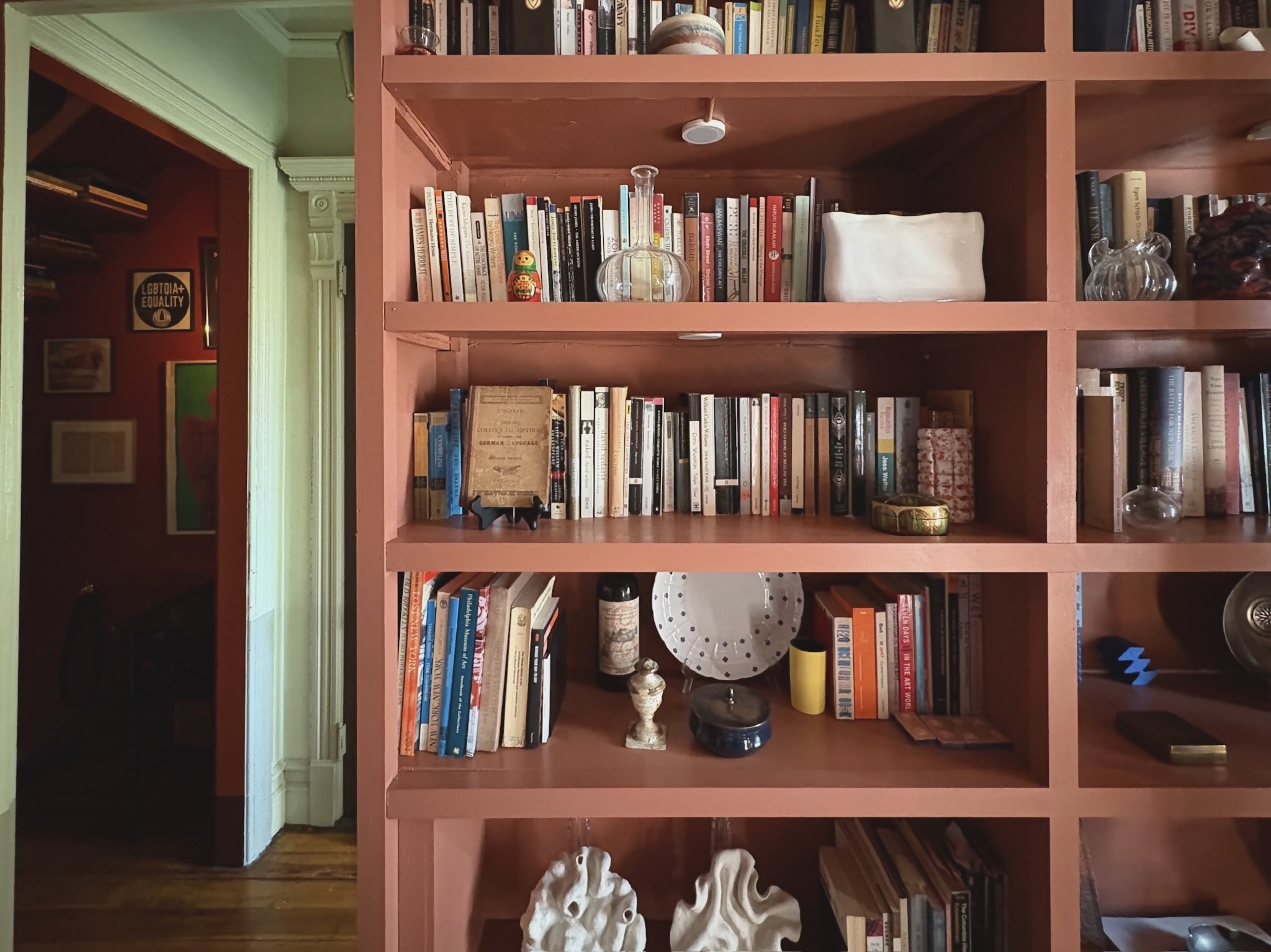 A corner of a room with a pink bookshelf filled with books, decorative objects, and art pieces. Part of a doorway with a green wall visible on the side.