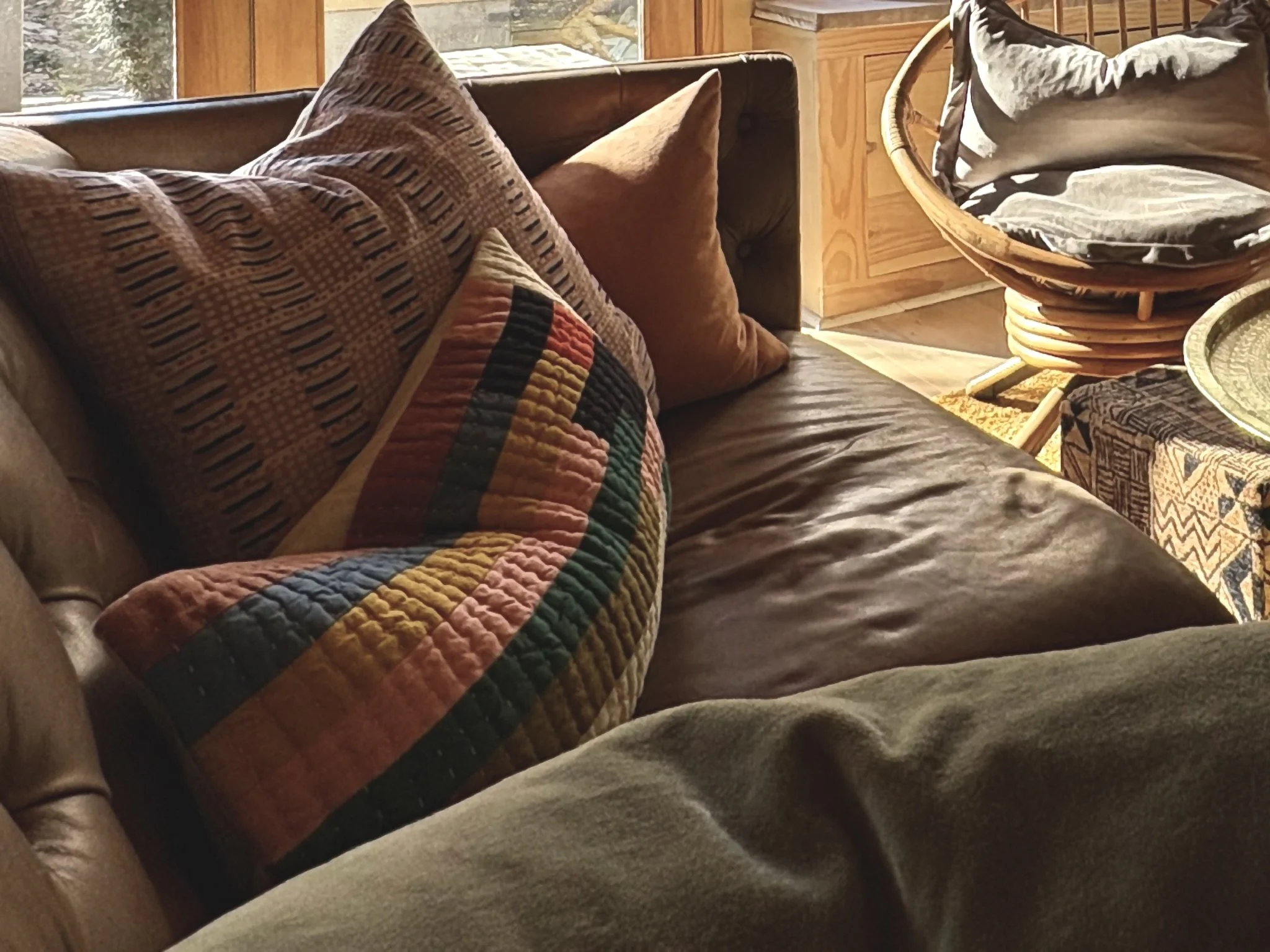 A cozy living room with a leather sofa adorned with patterned and plain throw pillows. Sunlight streams in from a window, highlighting the wooden furniture and a rattan armchair with cushions nearby.