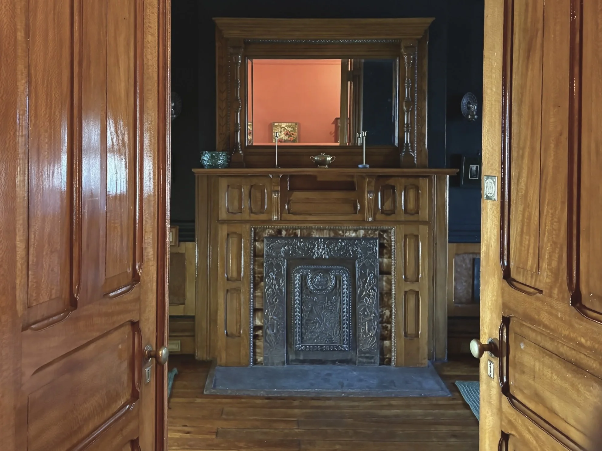 View through doorways into a room with an ornate wooden fireplace mantel and a mirror on top of it.