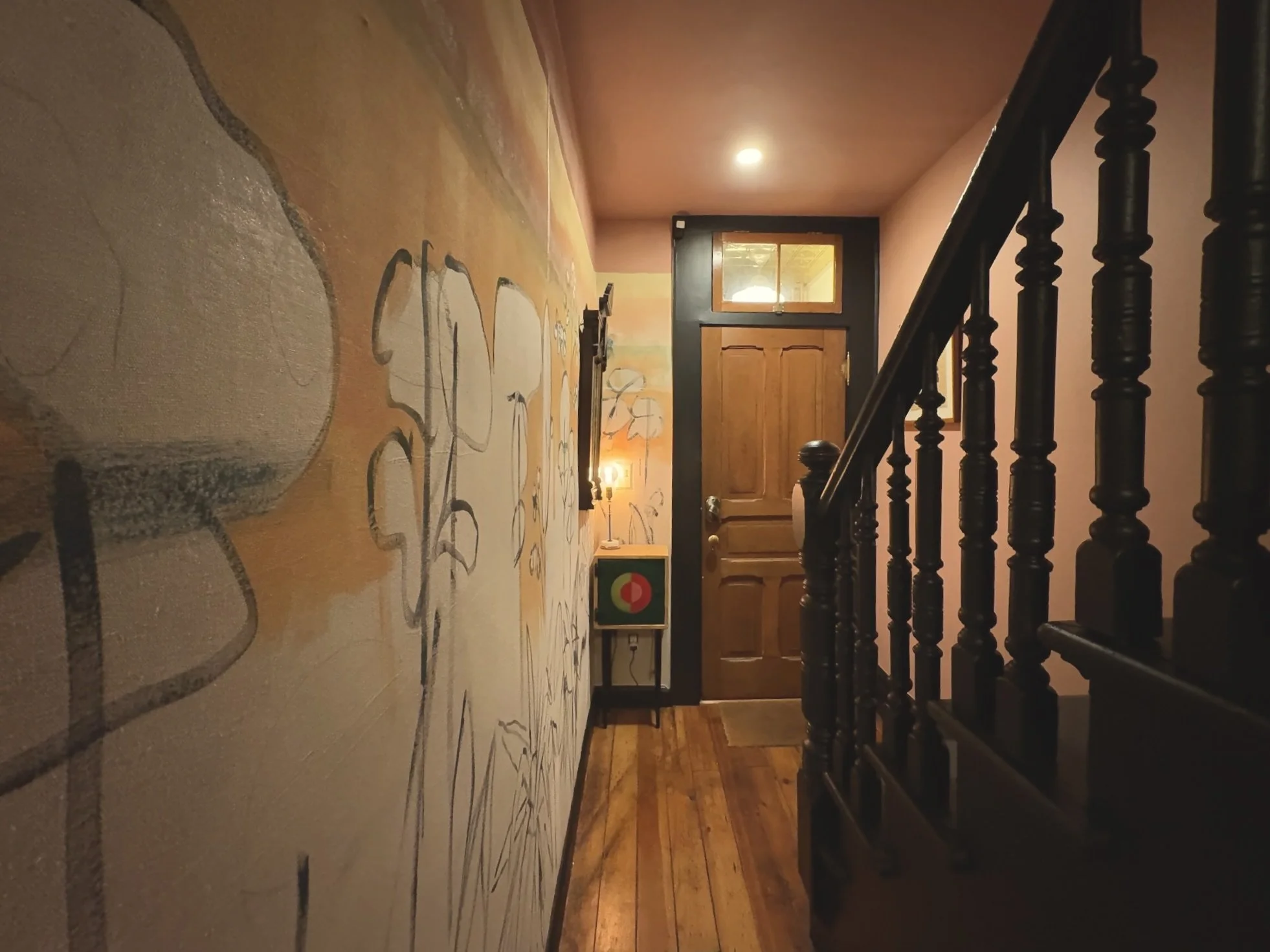 Interior view of a hallway with wooden flooring, a staircase with dark wood balusters to the right, and a closed wooden door at the end. A small table with a lamp and a colorful picture are visible near the door. The left wall has floral artwork and 