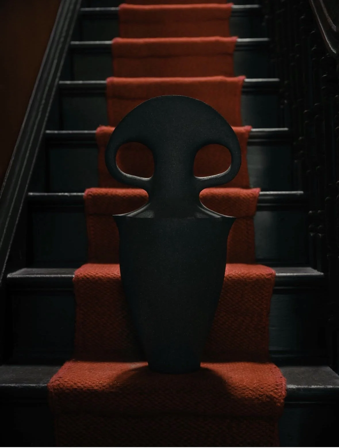A black, modern skull-shaped mask on a step with orange carpet, staircase with red carpeting behind.