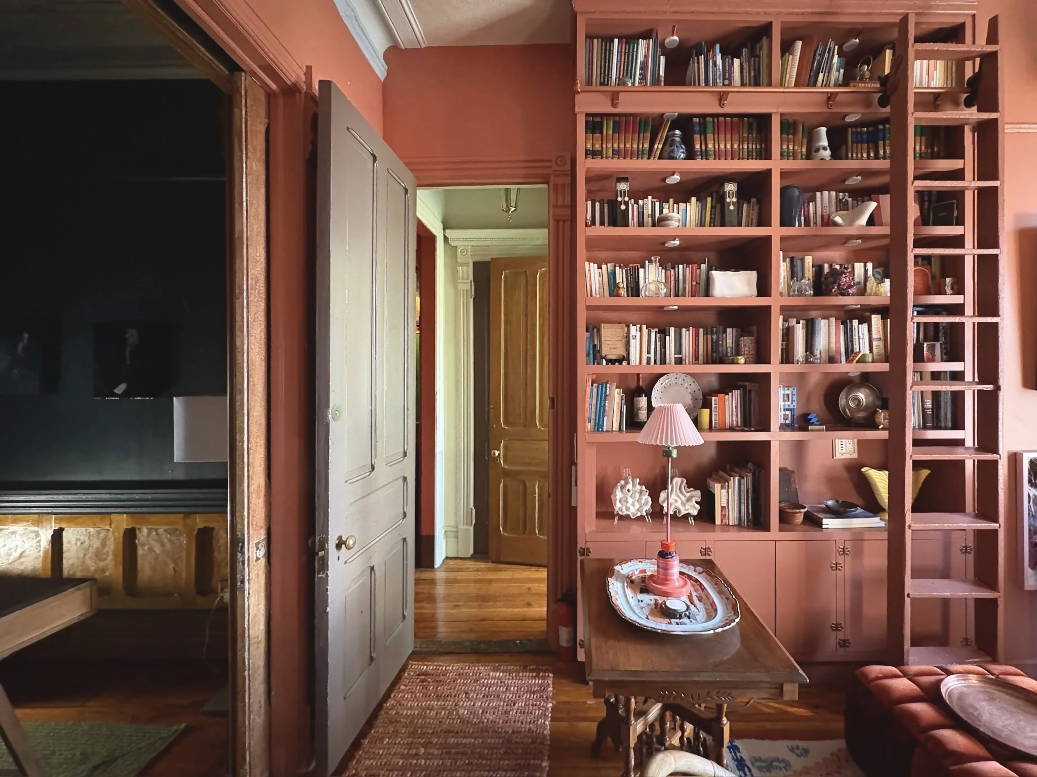 Interior view of a room with a pink wall and a large built-in bookshelf filled with books and decorative items. A small wooden table with a pink-lamp and tray sits in front of the bookshelf. An open door leads to a corridor with wooden floors and doo