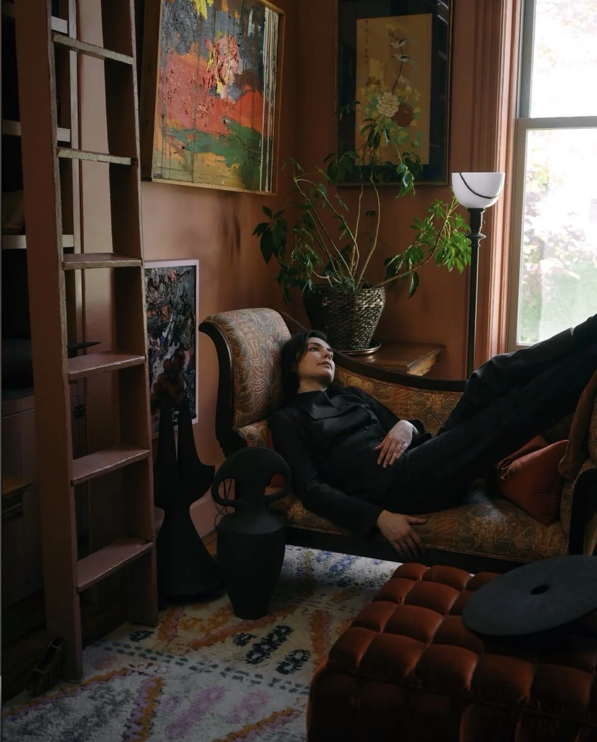 Woman lounging on a vintage sofa in a cozy, artistically decorated living room with paintings, indoor plants, a tall lamp, and a large window.
