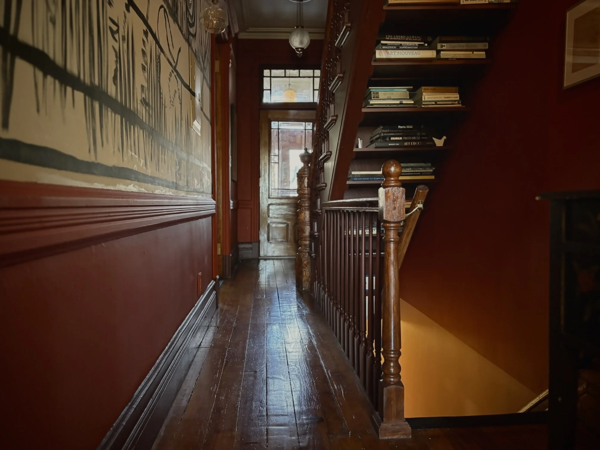 Wooden hallway with stairs and bookshelves leading to a door, decorated with antique Art Nouveau style wallpaper on the wall.