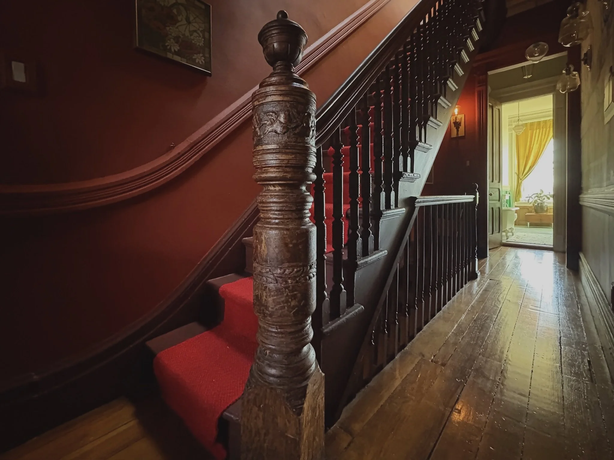 Interior view of a vintage wooden staircase with a red carpet runner leading to a brightly lit room with yellow curtains and furniture.